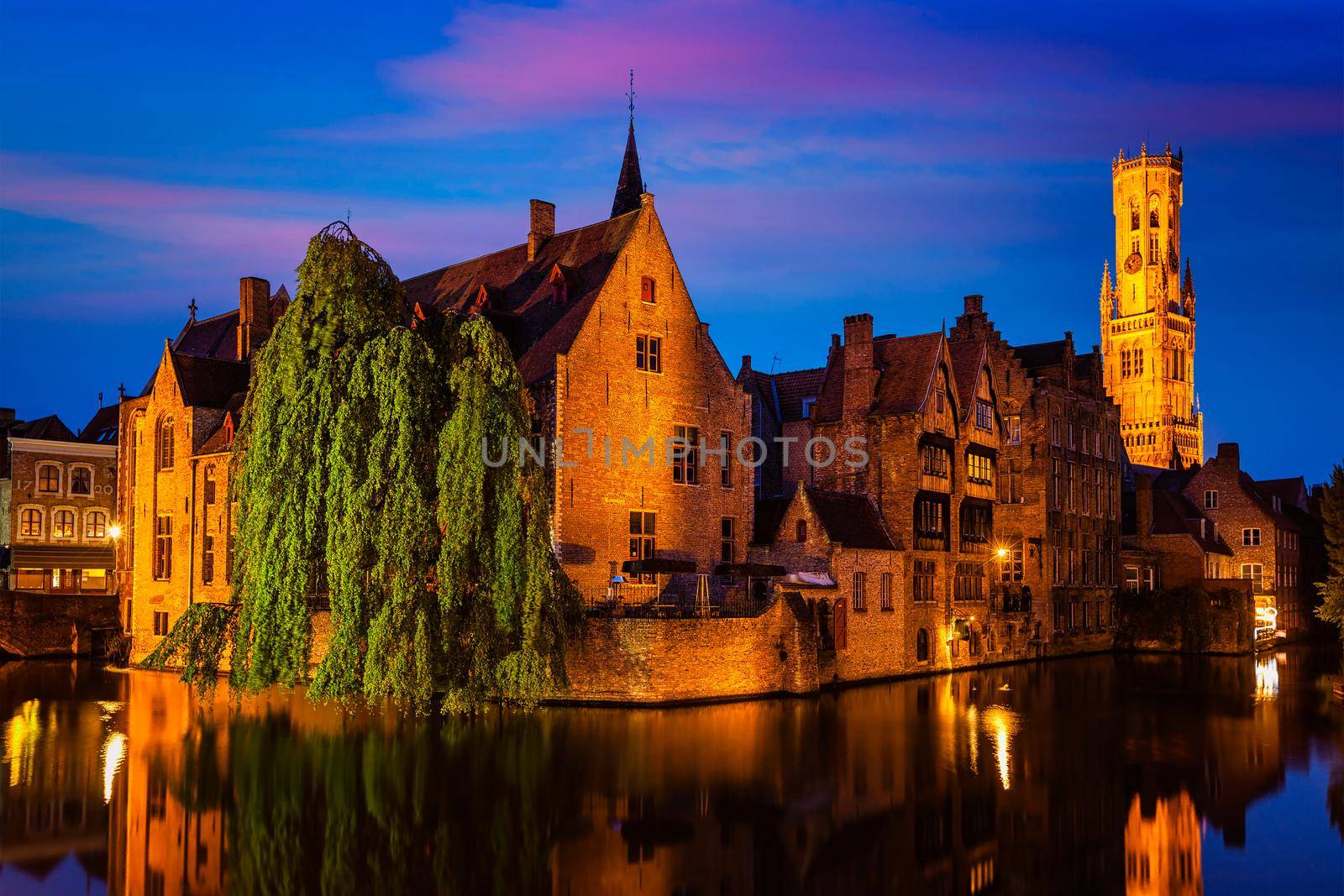 Famous view of Bruges - Rozenhoedkaai with Belfry and old houses along canal with tree in the night. Brugge, Belgium