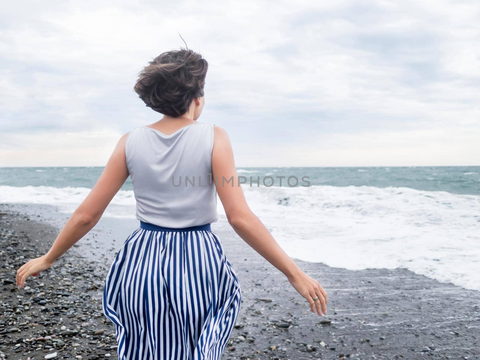 Happy woman greets stormy sea on pebble beach. Woman with hair ruffled with the wind. Wanderlust concept. Vacation on sea coast.