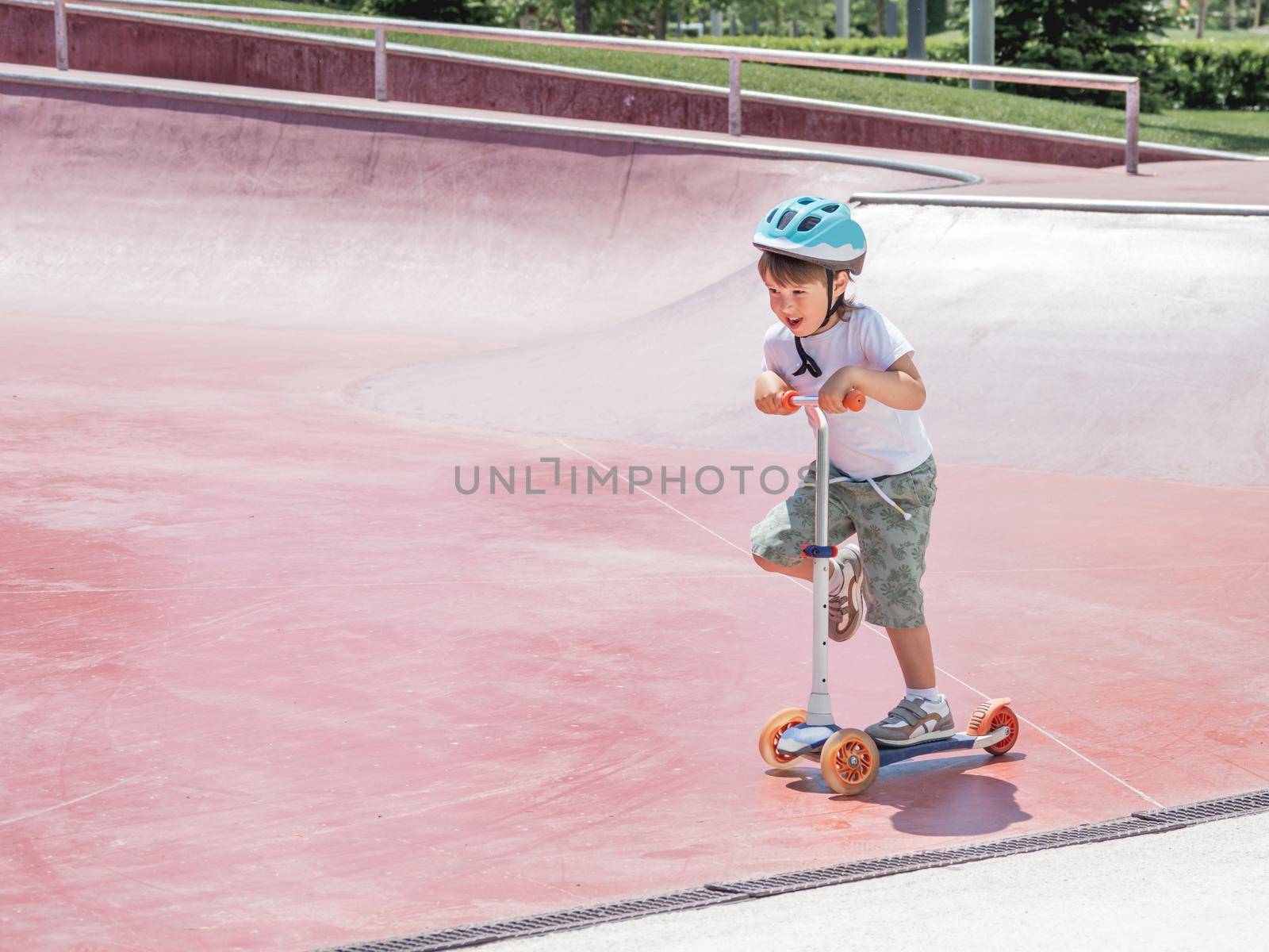 Little boy rides kick scooter in skate park. Special concrete bowl structures in urban park. Training to skate at summer. by aksenovko