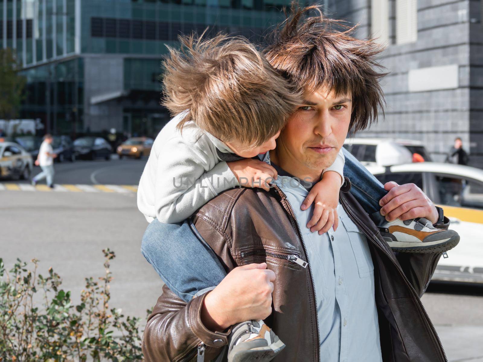 Little boy sits on father's shoulders between skyscrapers. Dad and son looks on glass walls of buildings. Future and modern technologies, life balance and family life in well keeps districts.