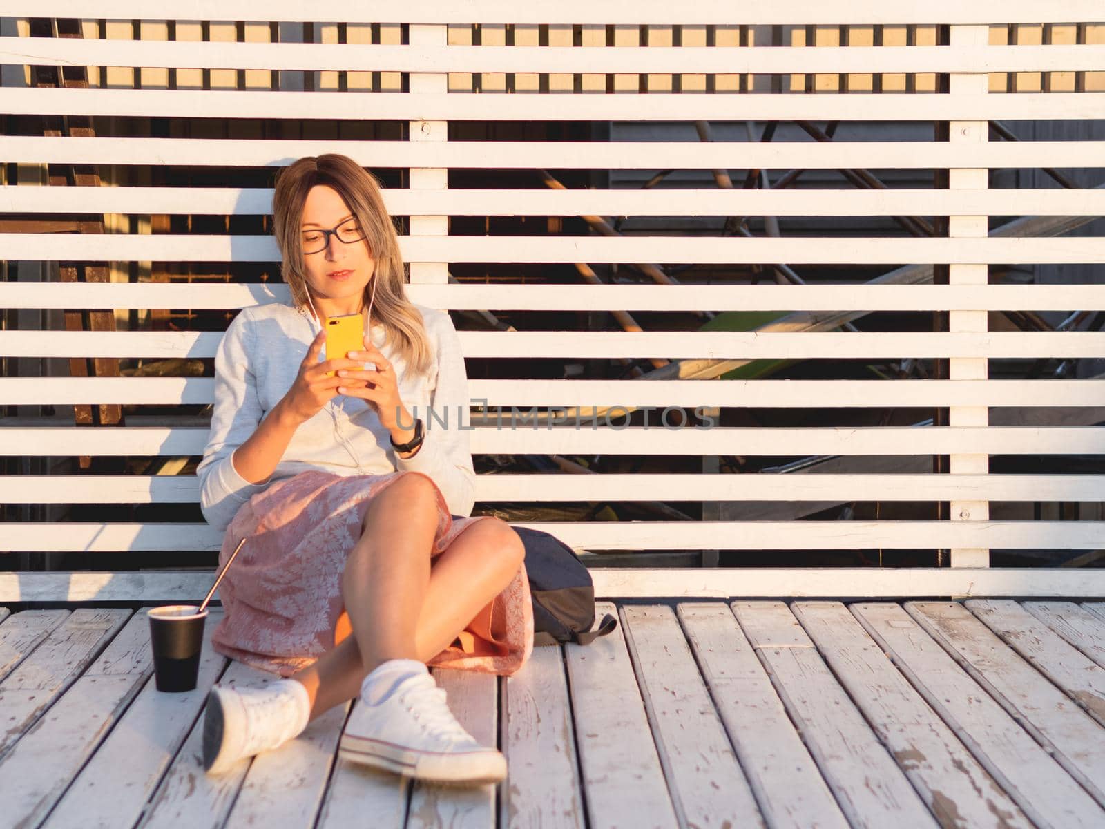 Woman with cup of coffee meets sunset on wooden pier. Female with curly hair and eyeglasses listens to music from smartphone. Enjoying nature outdoors. Summer casual clothes. Summer vibes.