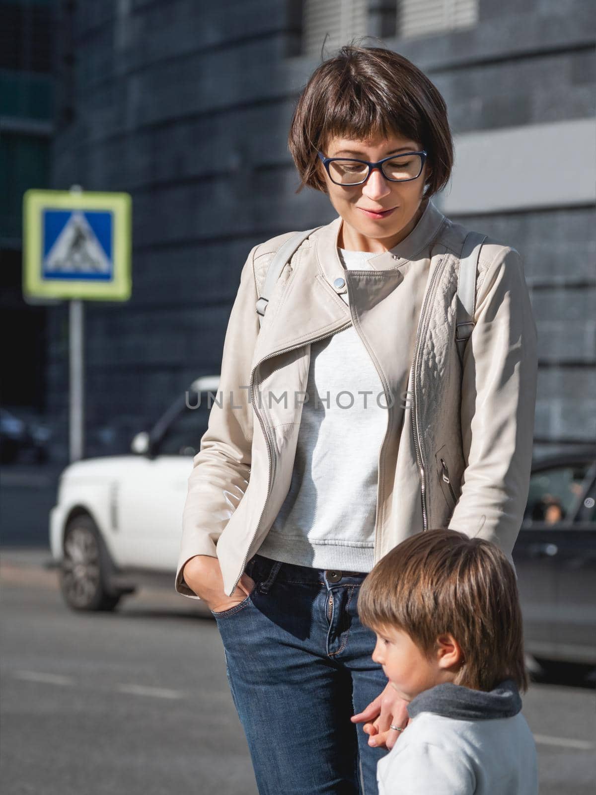 Mother and son stand on street in sunlight. Woman and kid in modern town with transport traffic and road sign. Family in city.