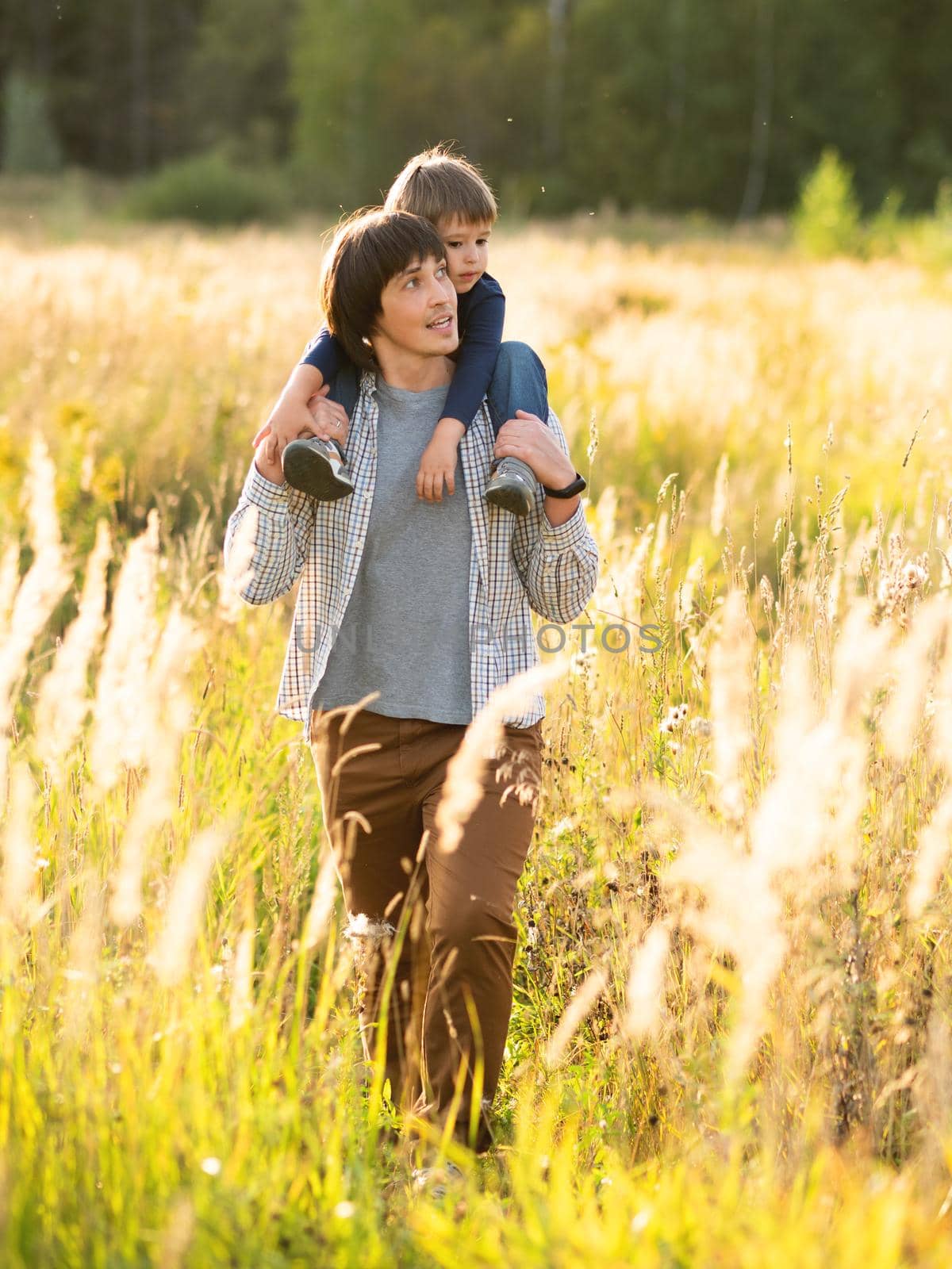 Father carries his son on shoulders. Family time outdoors. Man with child on autumn field at sunset. Dad and son explores nature together.
