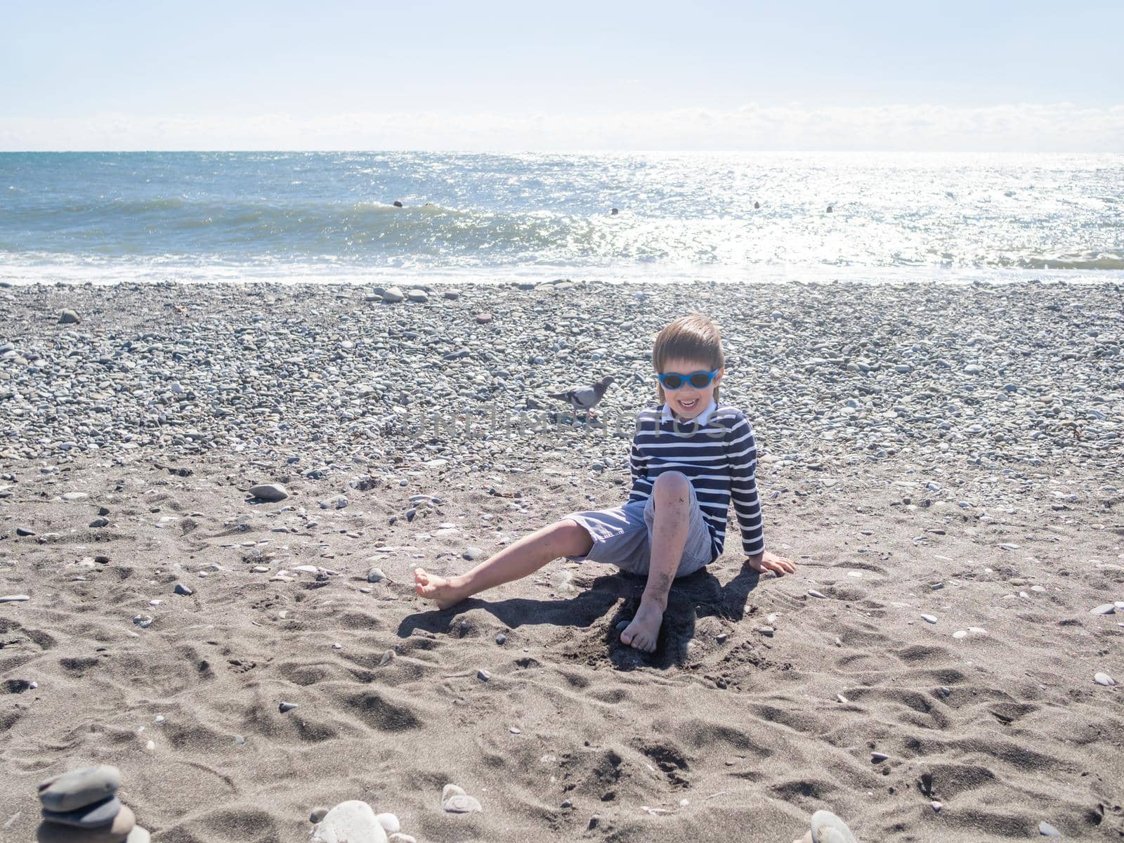 Happy boy is playing with wet sand, stones and driftwood on pebble beach. Vacation on seaside. Exploring nature in childhood. by aksenovko