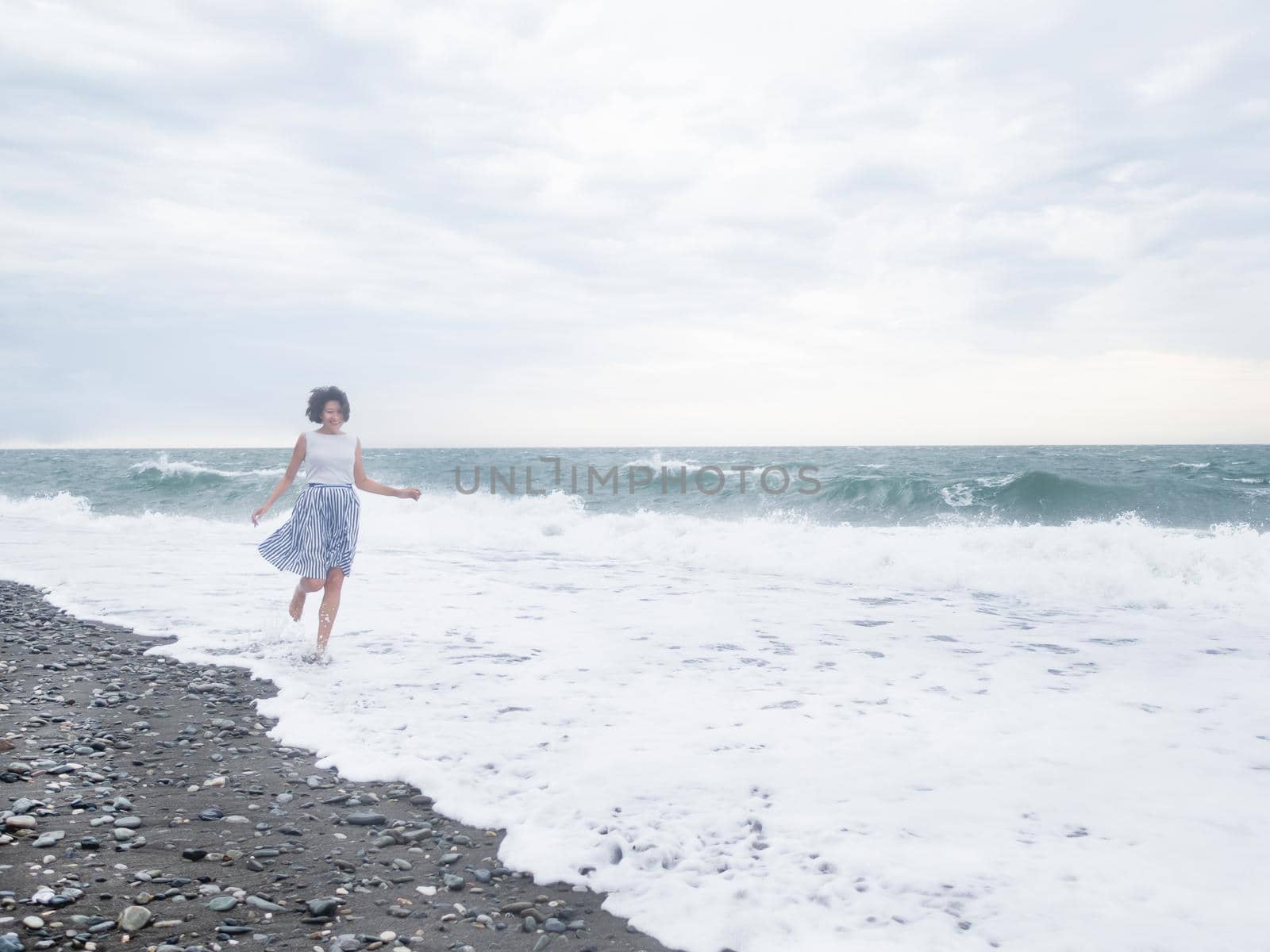 Happy woman is running on surf of stormy sea. Woman with hair ruffled with the wind. Wanderlust concept. Vacation on sea coast. by aksenovko