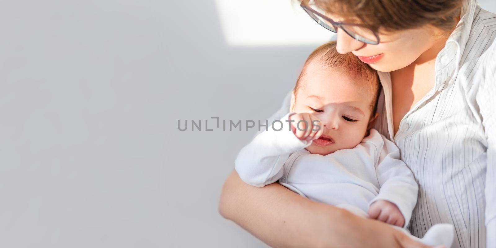 Mother hugs little baby in white onesie. Mom and 3-month old son or daughter. Family life. Horizontal banner with copy space.