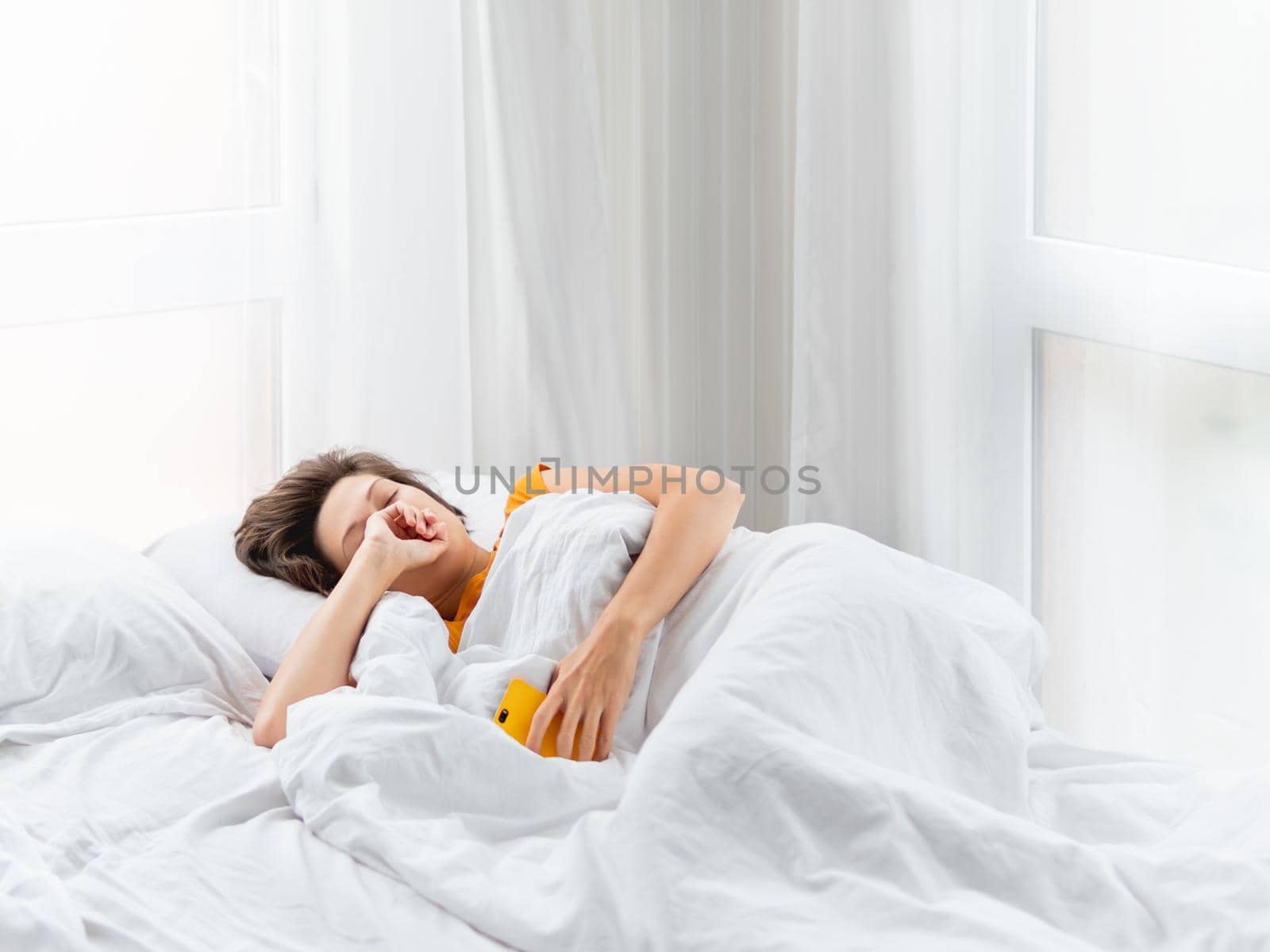 Sleepy woman is lying in bed, completely covered with white blanket. Smartphone used as alarm clock. It's hard to wake up early in morning. Woman does not get enough sleep. by aksenovko