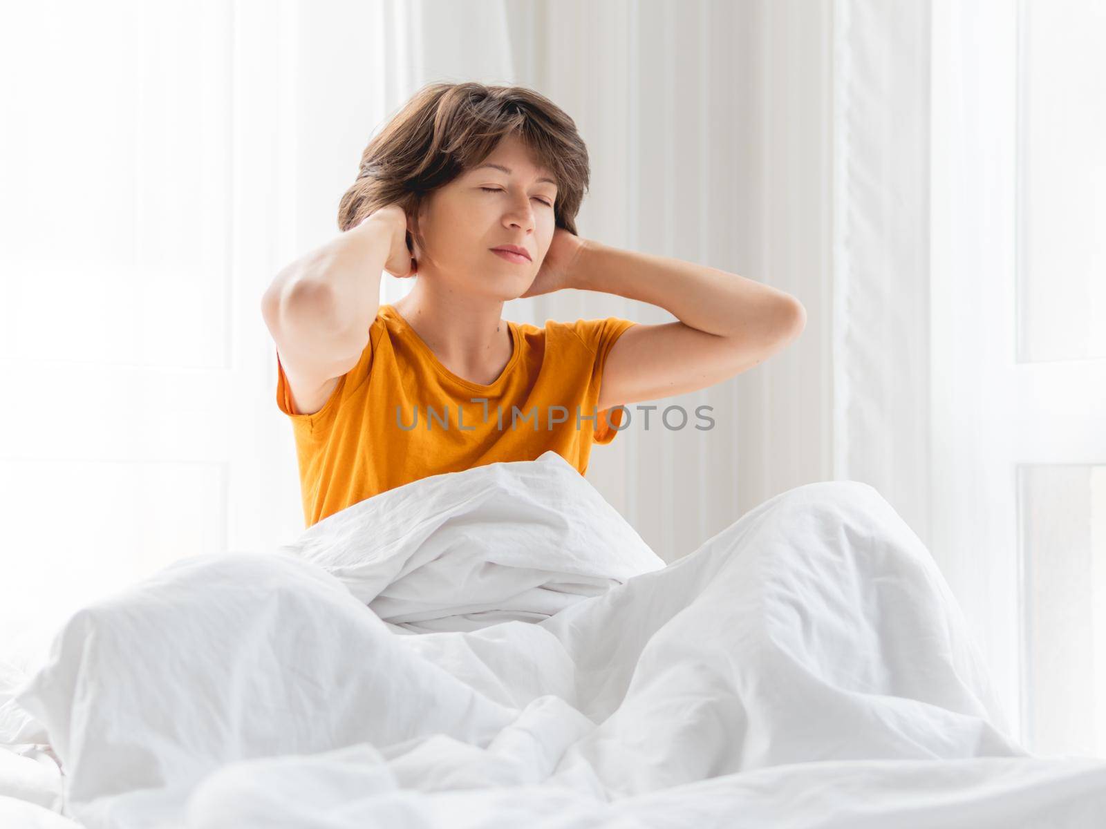 Sleepy woman in yellow pajama is massaging her neck in bed, covered with white blanket. Waking up early in morning.