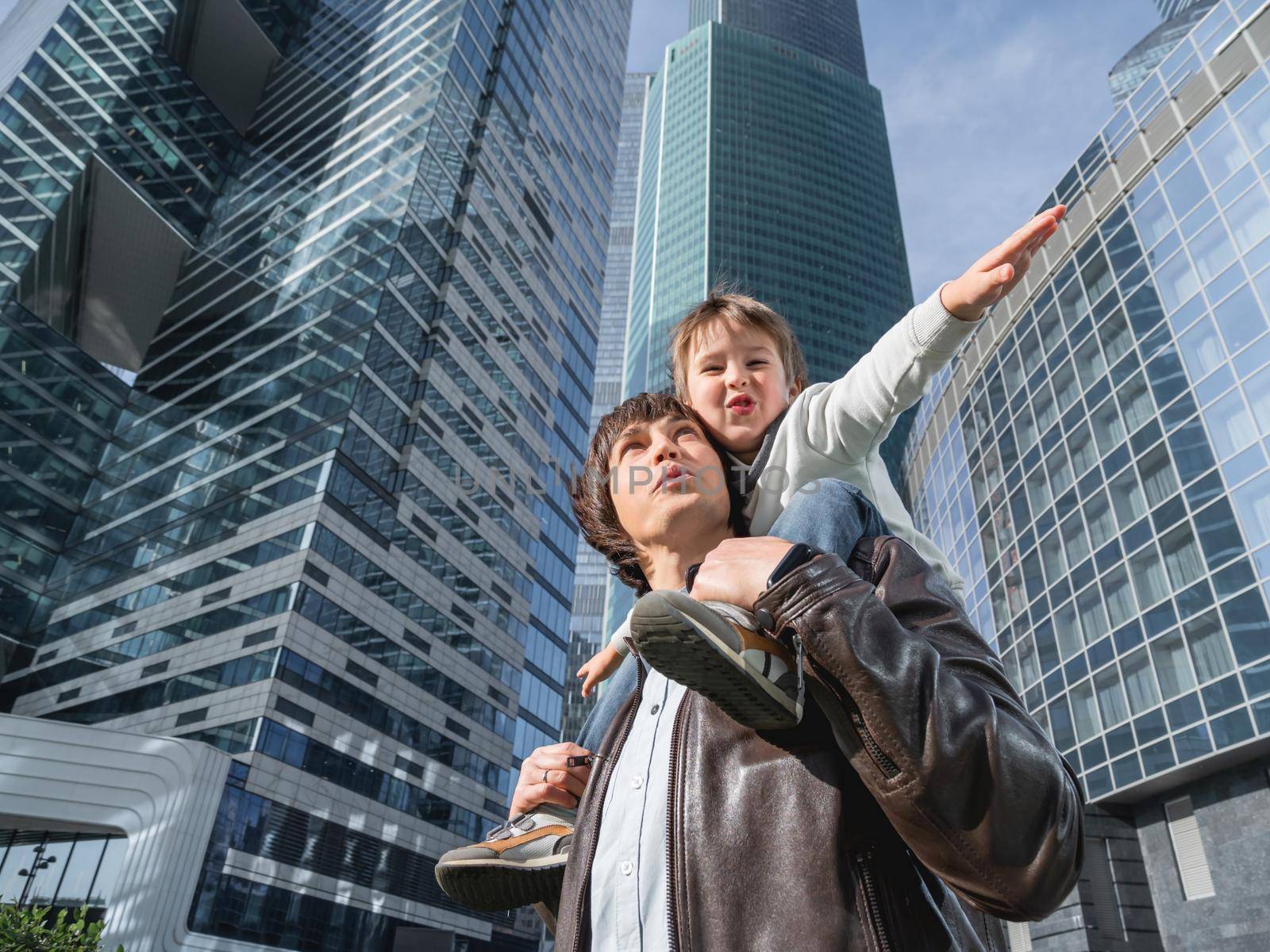 Laughing boy plays like airplane on father's shoulders. Dad and son looks on glass walls of buildings. Future and modern technologies, life balance and family life in well keeps districts.