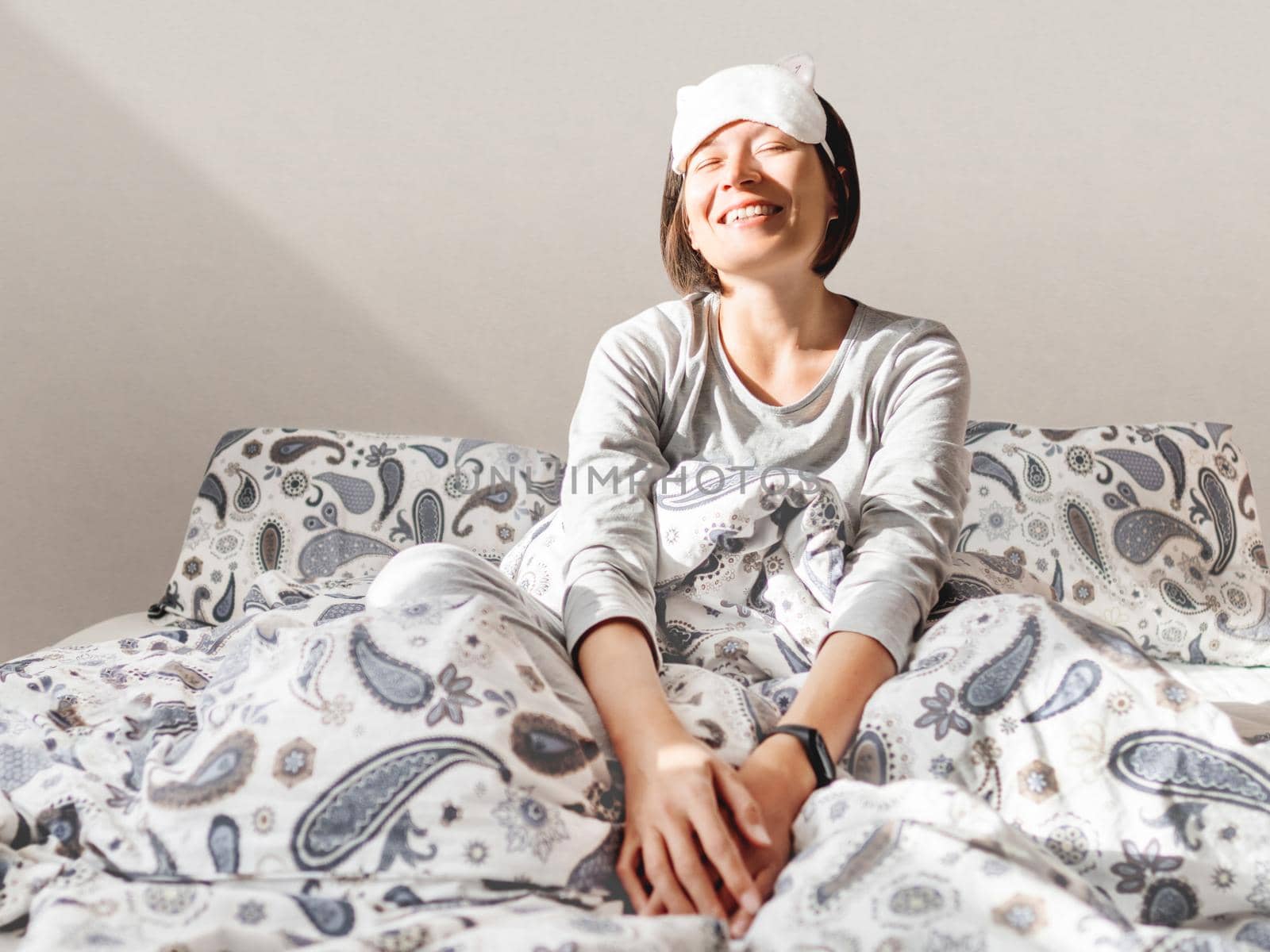 Smiling woman in grey pajama and sleeping mask. She is just woke up and sit in bed. Early morning in cozy home. by aksenovko