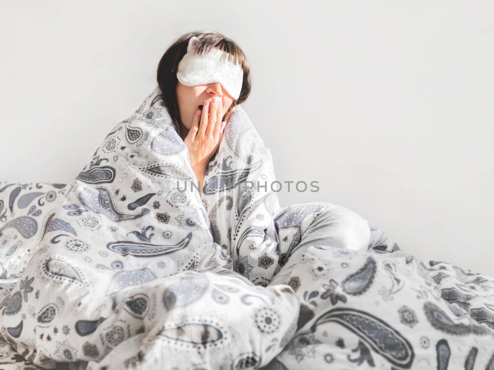 Woman in grey pajama and sleeping mask on face. She is just woke up and sit in bed. Early morning in cozy home.