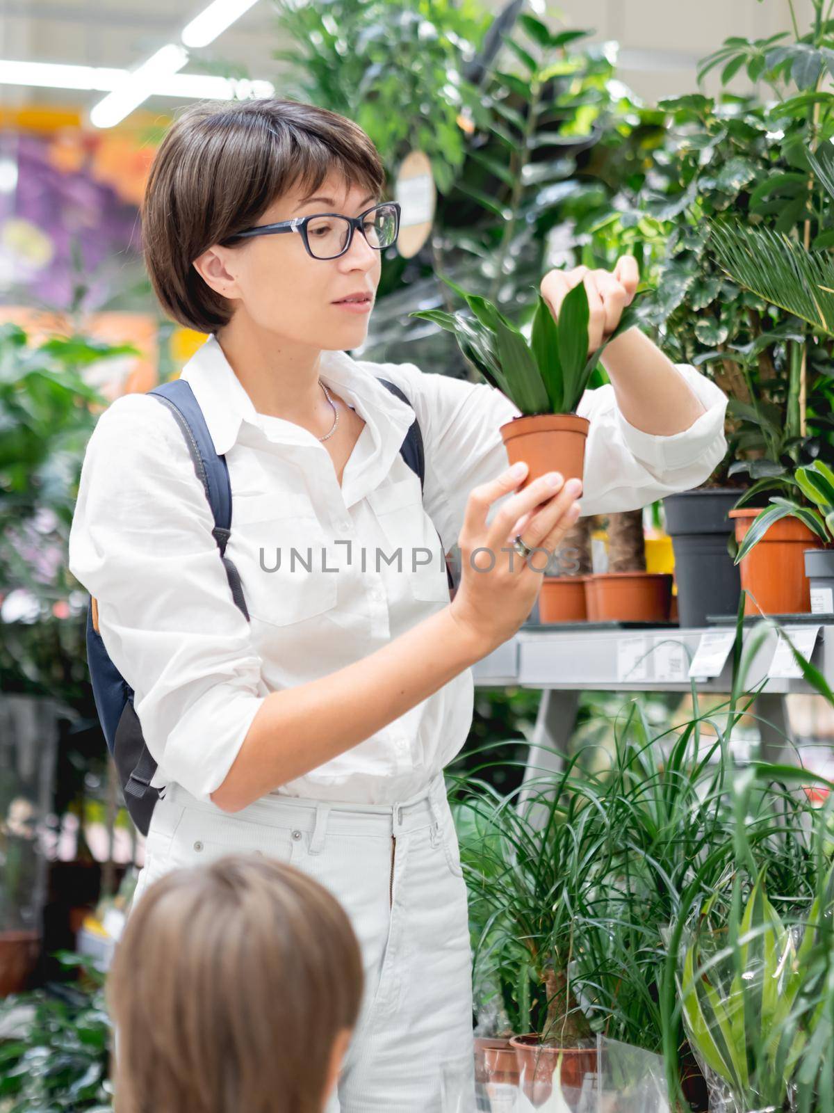 Woman and kid choose plants for home. Mother and son near shelves with seedlings, flowering plants and seeds in flower shop. Indoor agronomic market. by aksenovko