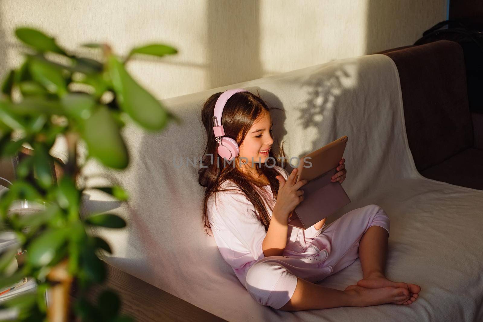 A little adorable girl in pink home clothes and pink headphones sits on the couch, looks into a digital tablet.