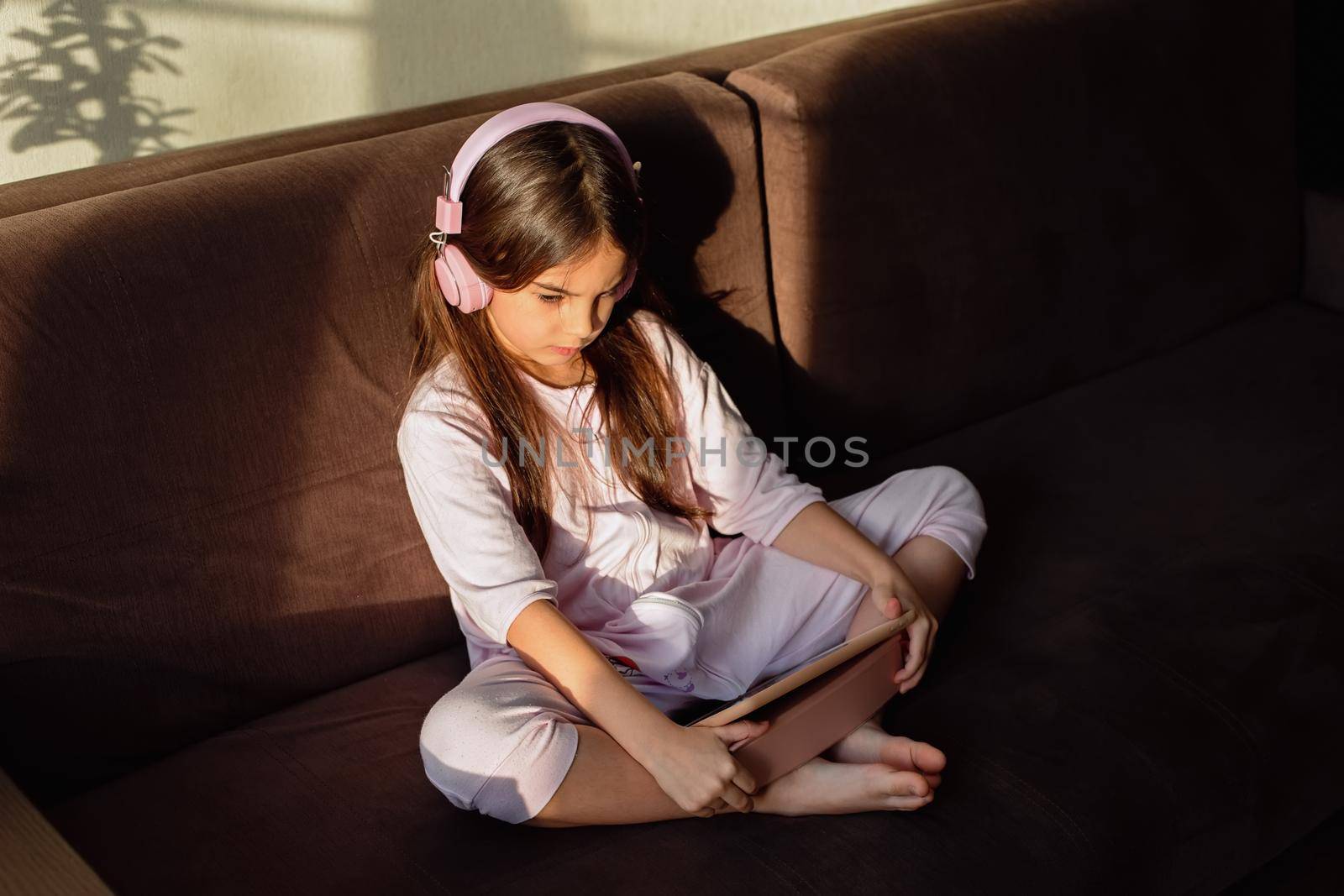 A brunette little girl in pink headphones sits on the couch, looks into a digital tablet. by Zakharova