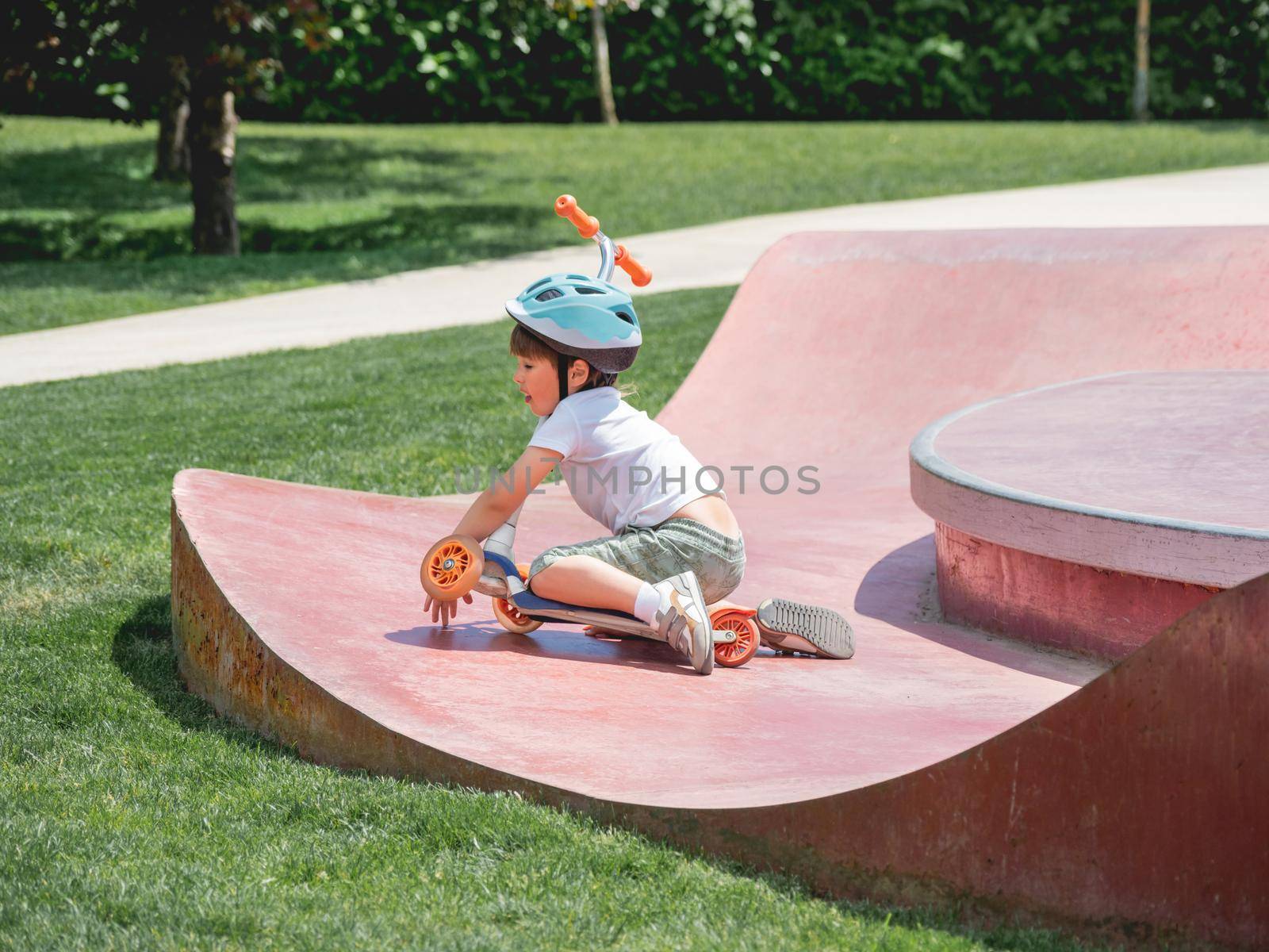 Little boy fell off kick scooter while riding in skate park. Special concrete bowl structures in urban park. Training to skate at summer.