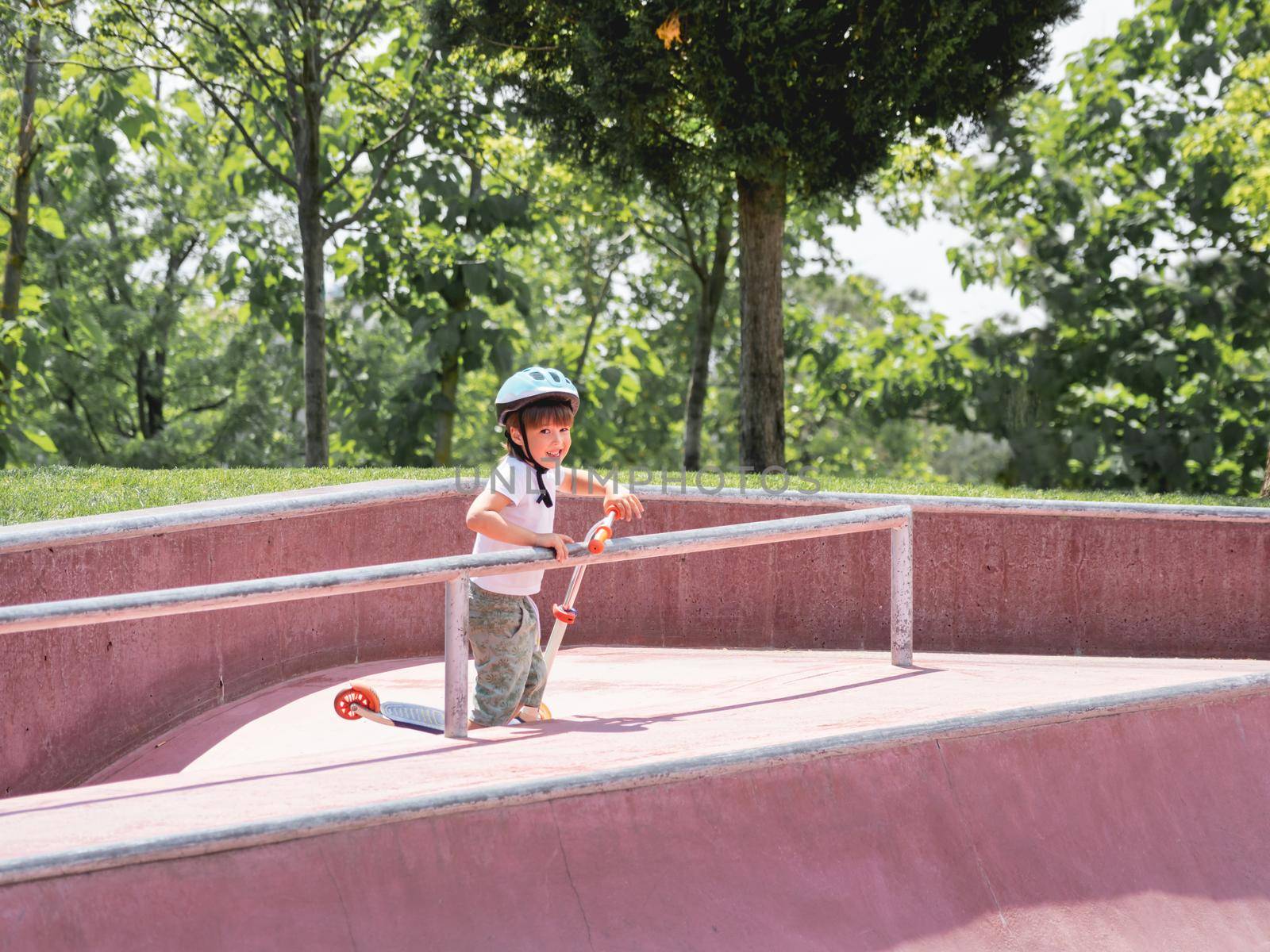 Little boy rides kick scooter in skate park. Special concrete bowl structures in urban park. Training to skate at summer.