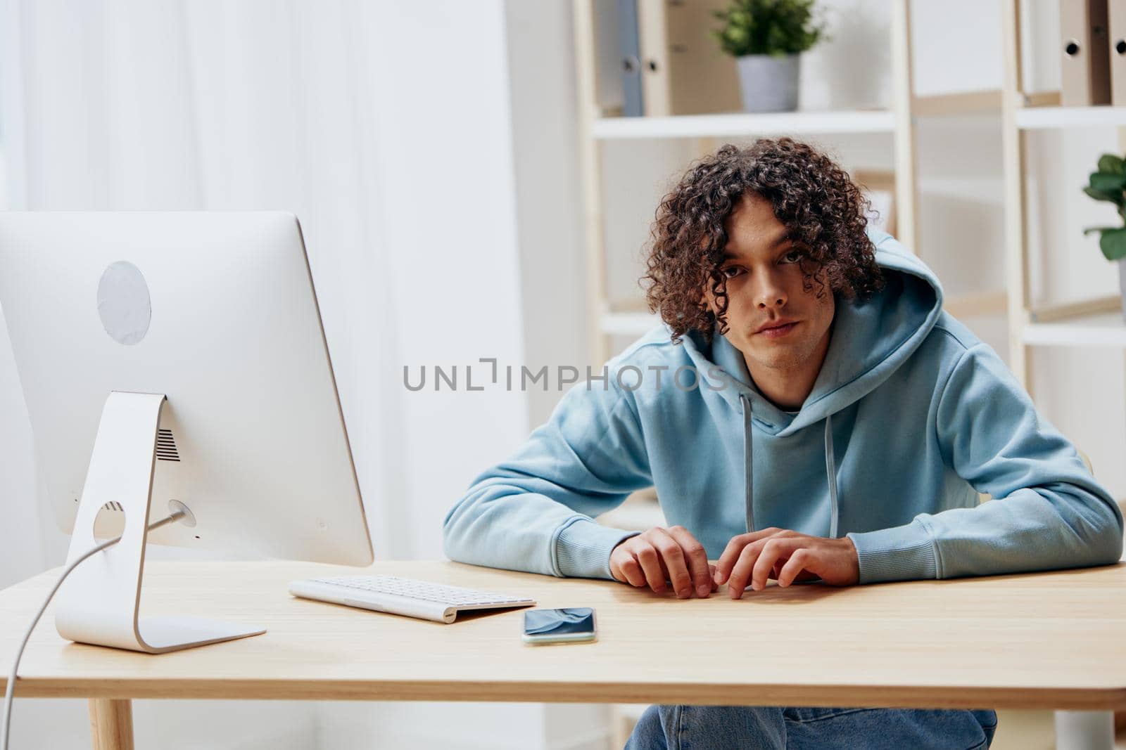 A young man in a blue jacket in front of a computer with phone Lifestyle by SHOTPRIME