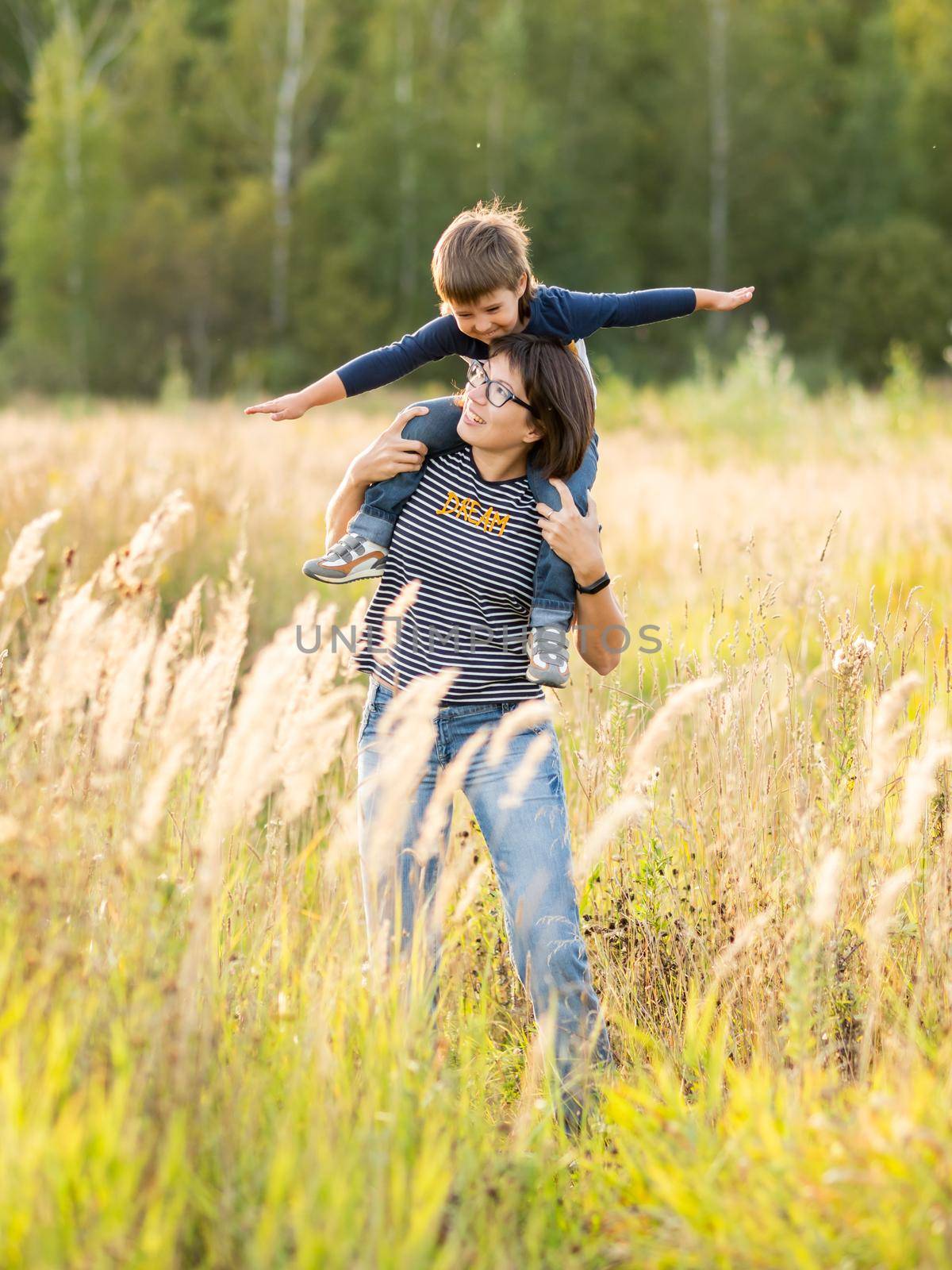 Cute boy and his mother play as airplane. Happy kid dreams to be a pilot. Boy is planning for the future. Mom and son on field at golden sunset hour at autumn season. by aksenovko