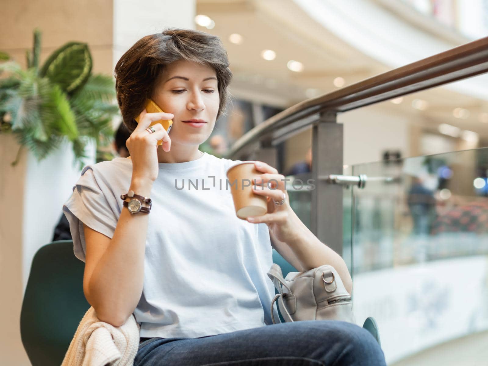 Woman is talking by smartphone and drinking coffee at shopping mall. Coffee break at store.