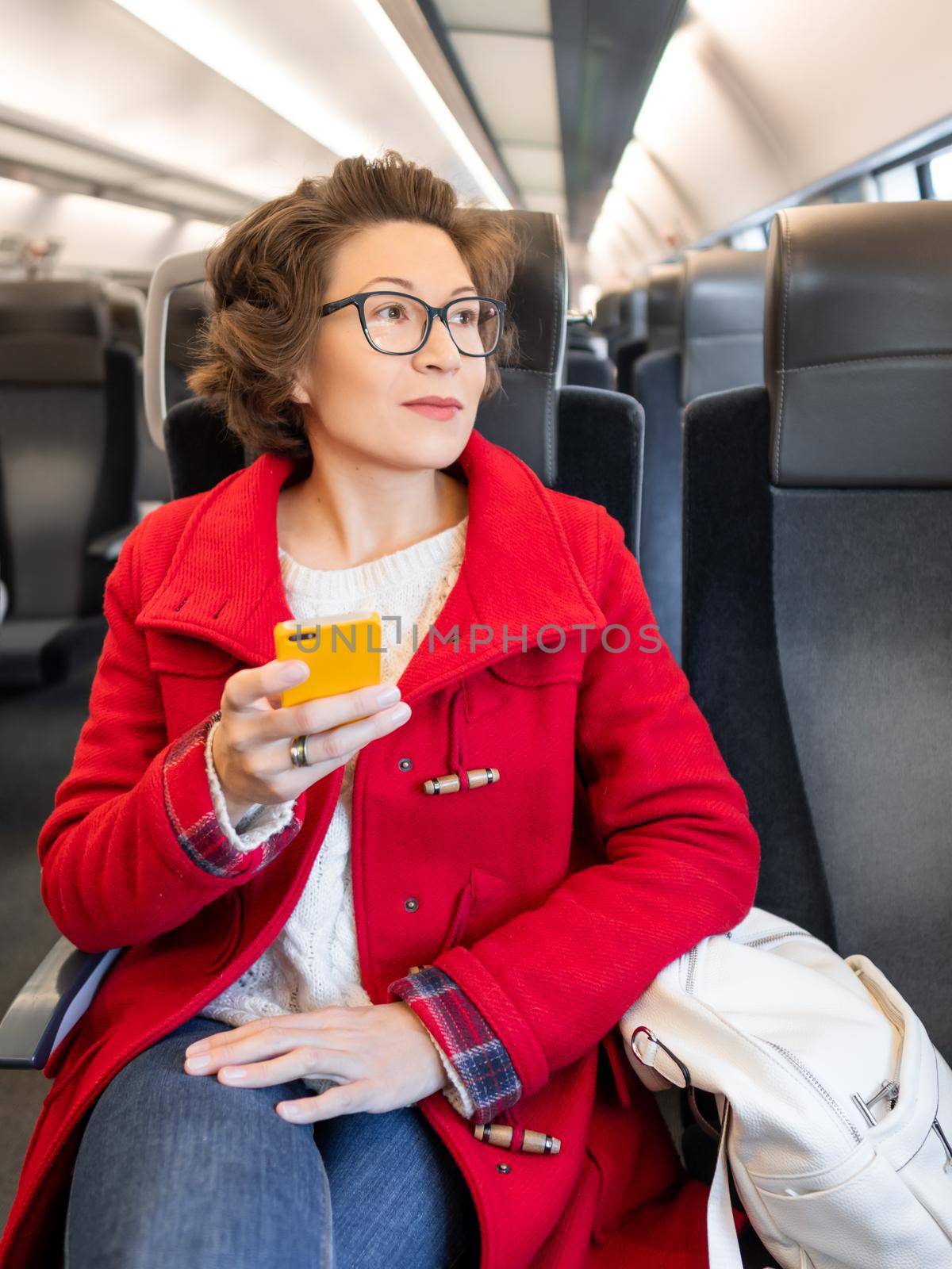 Woman in red duffle coat texting on smartphone in suburban train. Travel by land vehicle.