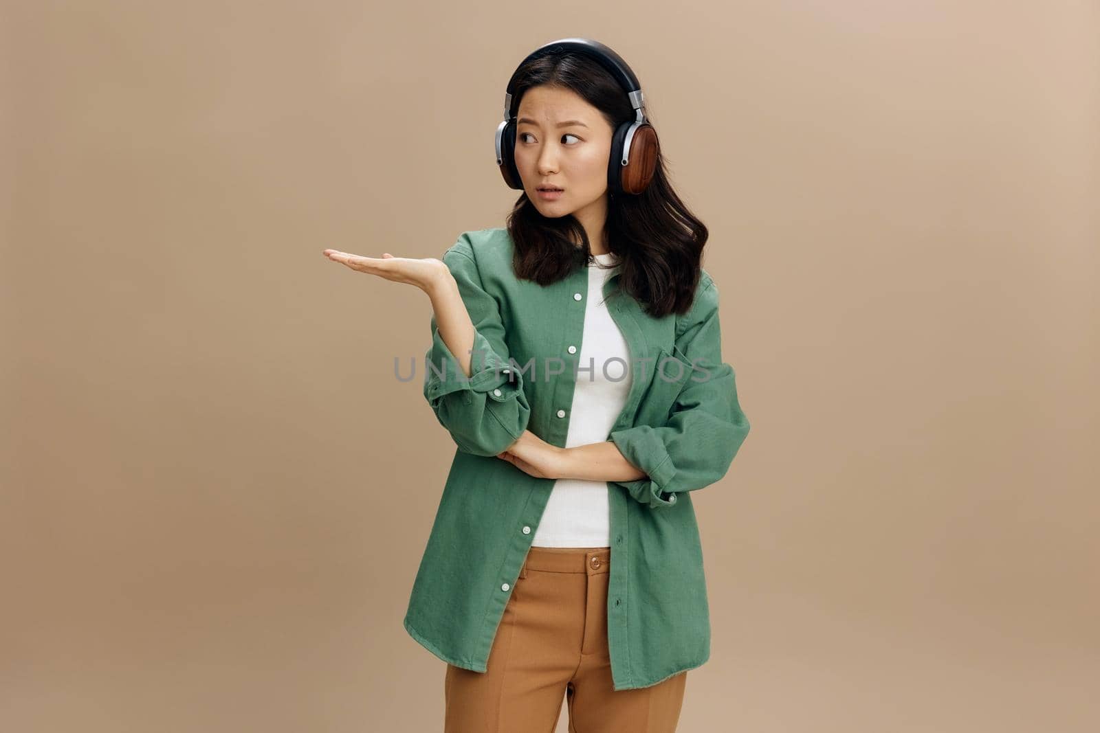 Wow, look here. Korean shocked young woman in khaki green shirt headphones hold palm up aside posing isolated on beige pastel studio background. Cool fashion offer. Music App Platform Ad concept
