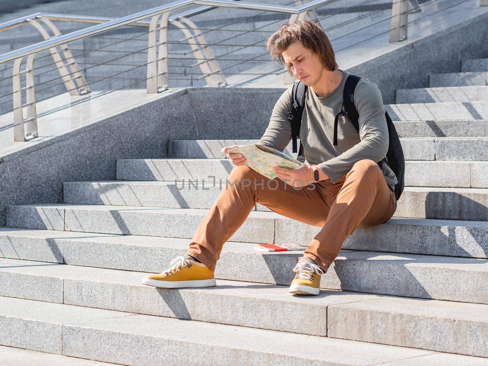 Man sits on stone staircase and reads map. Solo-travelling around city. Urban tourism. Modern architectural landmarks.