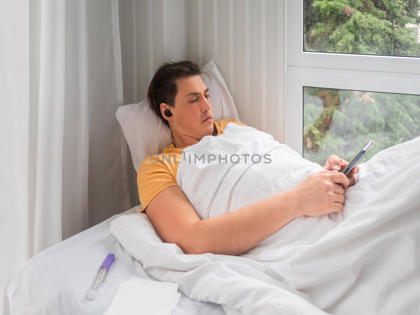 Sick man is lying in bed with thermometer. Man is searching online for disease symptoms or texting to his doctor. Using wireless technology in case of temporary disability. by aksenovko