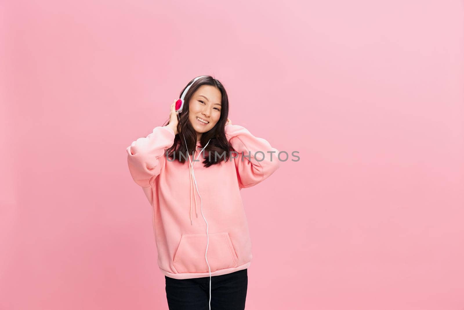 Enjoying Asian student young lady in pink hoodie sweatshirt with cute headphones smile touch head posing isolated on over pink studio background. Good offer. Sound streaming platform ad concept