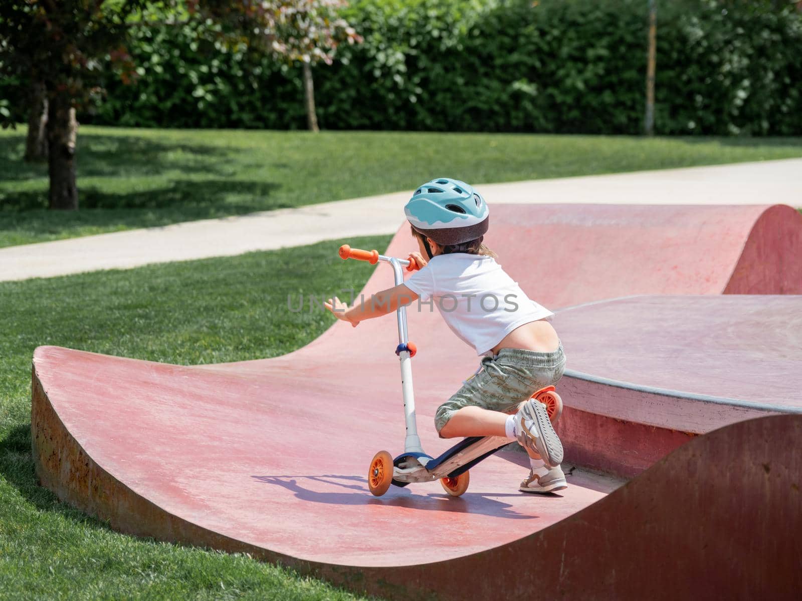 Little boy fell off kick scooter while riding in skate park. Special concrete bowl structures in urban park. Training to skate at summer. by aksenovko