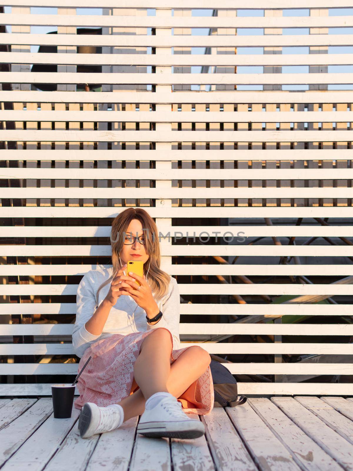 Woman with cup of coffee meets sunset on wooden pier. Female with curly hair and eyeglasses listens to music from smartphone. Enjoying nature outdoors. Summer casual clothes. Summer vibes. by aksenovko