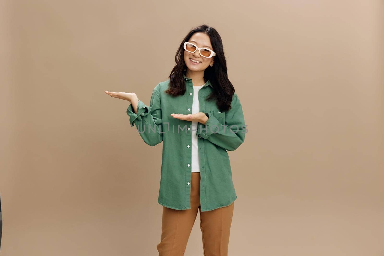 Hey, great purpose. Cheerful Korean woman in khaki green shirt stylish eyewear hold invisible objects on palms posing isolated on beige pastel background. Cool fashion offer. Sunglasses ad concept