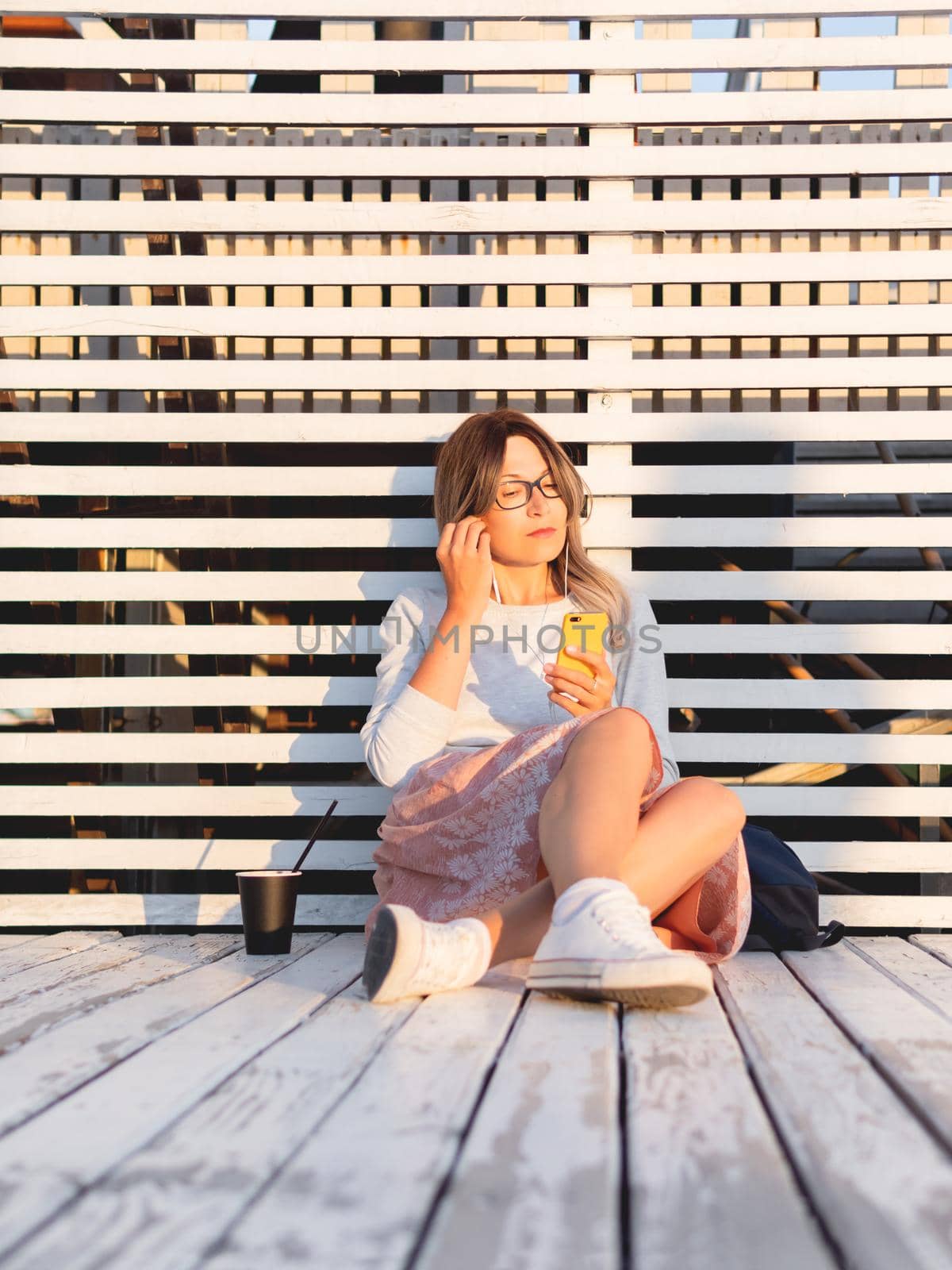 Woman with cup of coffee meets sunset on wooden pier. Female with curly hair and eyeglasses listens to music from smartphone. Enjoying nature outdoors. Summer casual clothes. Summer vibes. by aksenovko