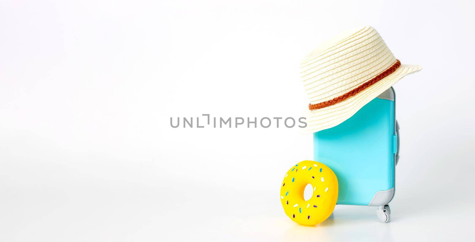 A blue suitcase on wheels, a straw hat, and a yellow inflatable donut stand on a white background. copy space. Wide banner