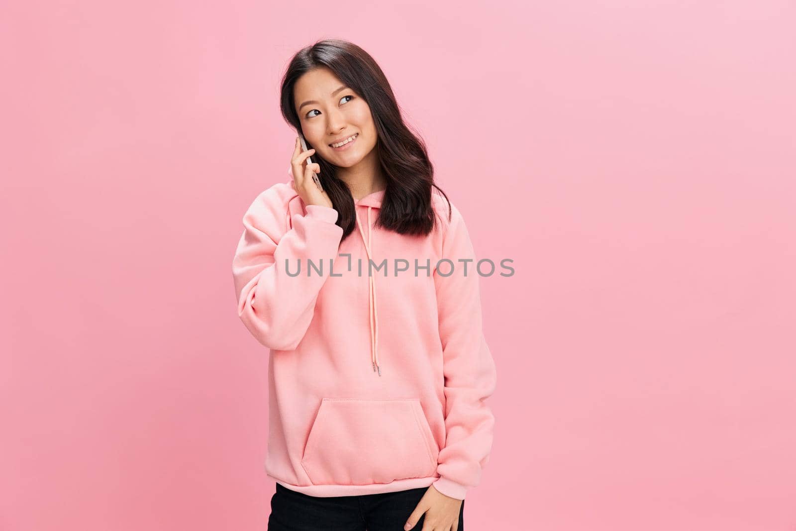 Love at a distance. Lovely happy Asian young lady in pink hoodie sweatshirt talk with boyfriend using phone posing isolated on pink studio background. Good offer. Gadget addiction Social Media concept