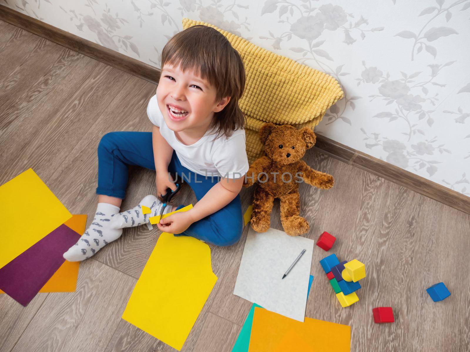 Toddler boy learns to cut colored paper with scissors. Kid sits on floor in kids room with toy blocks and teddy bear. Educational classes for children. Developing feeling sensations and fine motor skills at home. by aksenovko