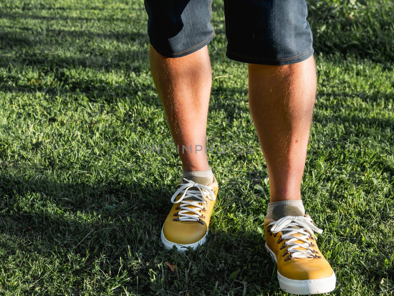 Man in bright yellow sneakers is standing on green grass lawn in park. Modern hipster's shoes. Urban fashion.