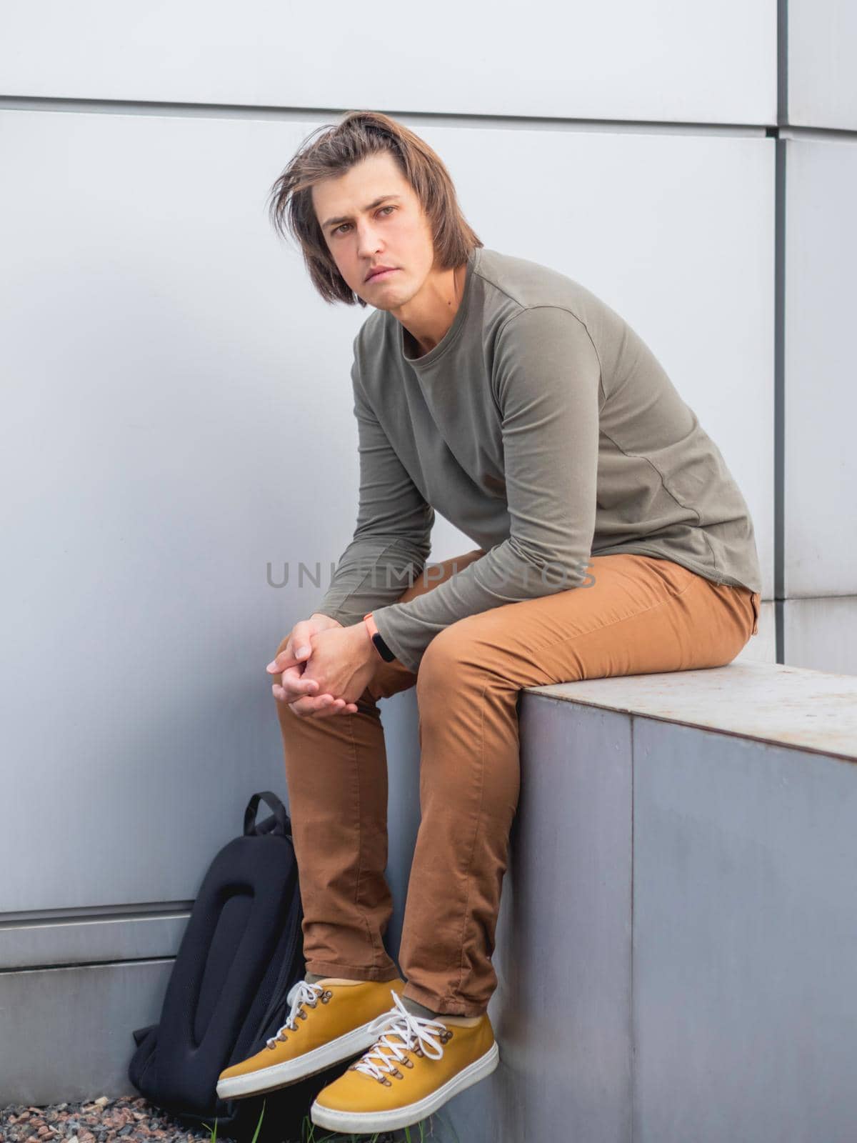 Millennial man with long hair and tense look sits near grey wall. Serious man in earth toned casual clothes is staring in camera.