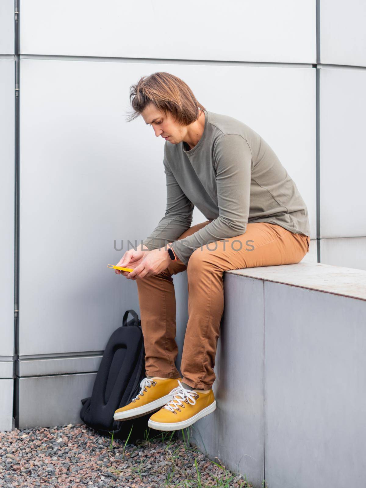 Thoughtful young man sits with smartphone on outdoor part of wall. Student with rucksack in casual clothes. Handsome man with long hair. Modern urban lifestyle.