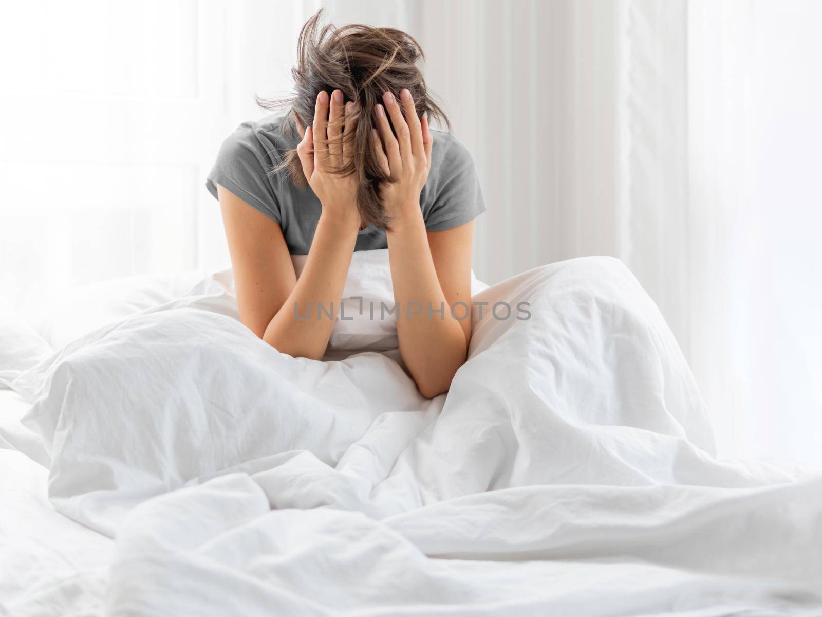 Sleepy woman with hands in hair is sitting in bed . Woman in depression or suffer of headache.