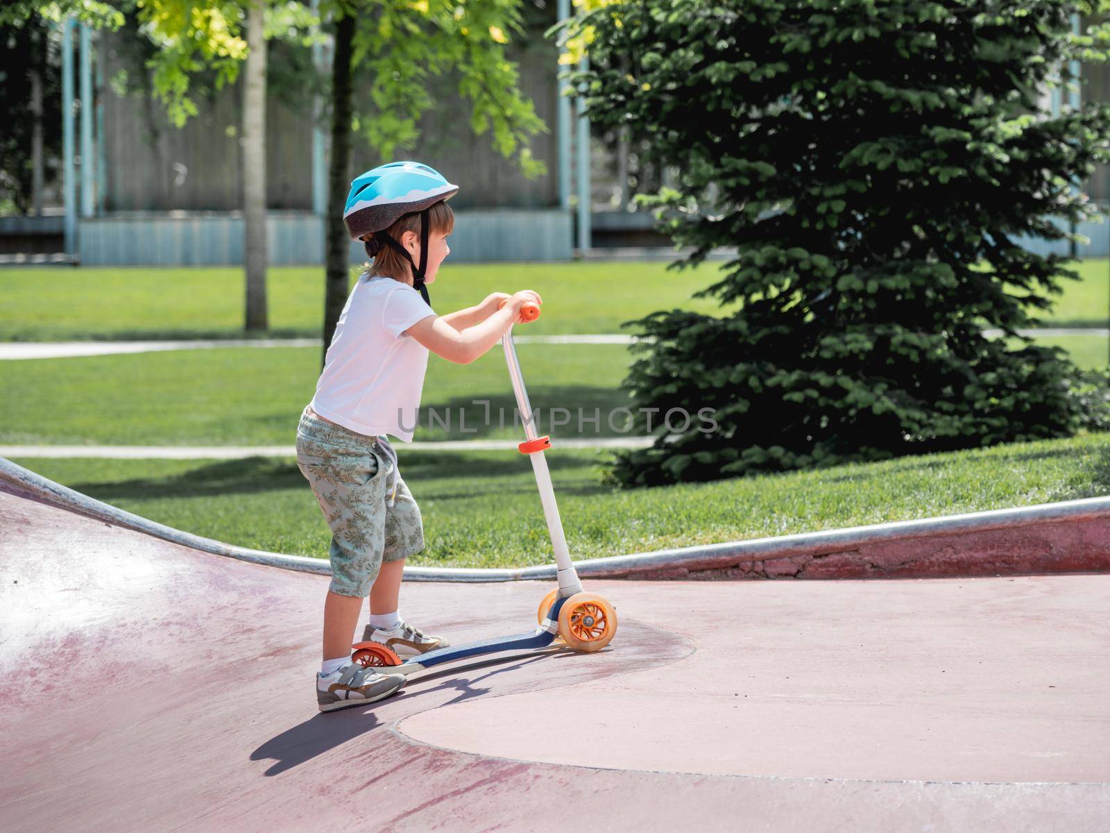 Brave boy rides kick scooter in skate park. Special concrete bowl structures in urban park. Training to skate at summer.