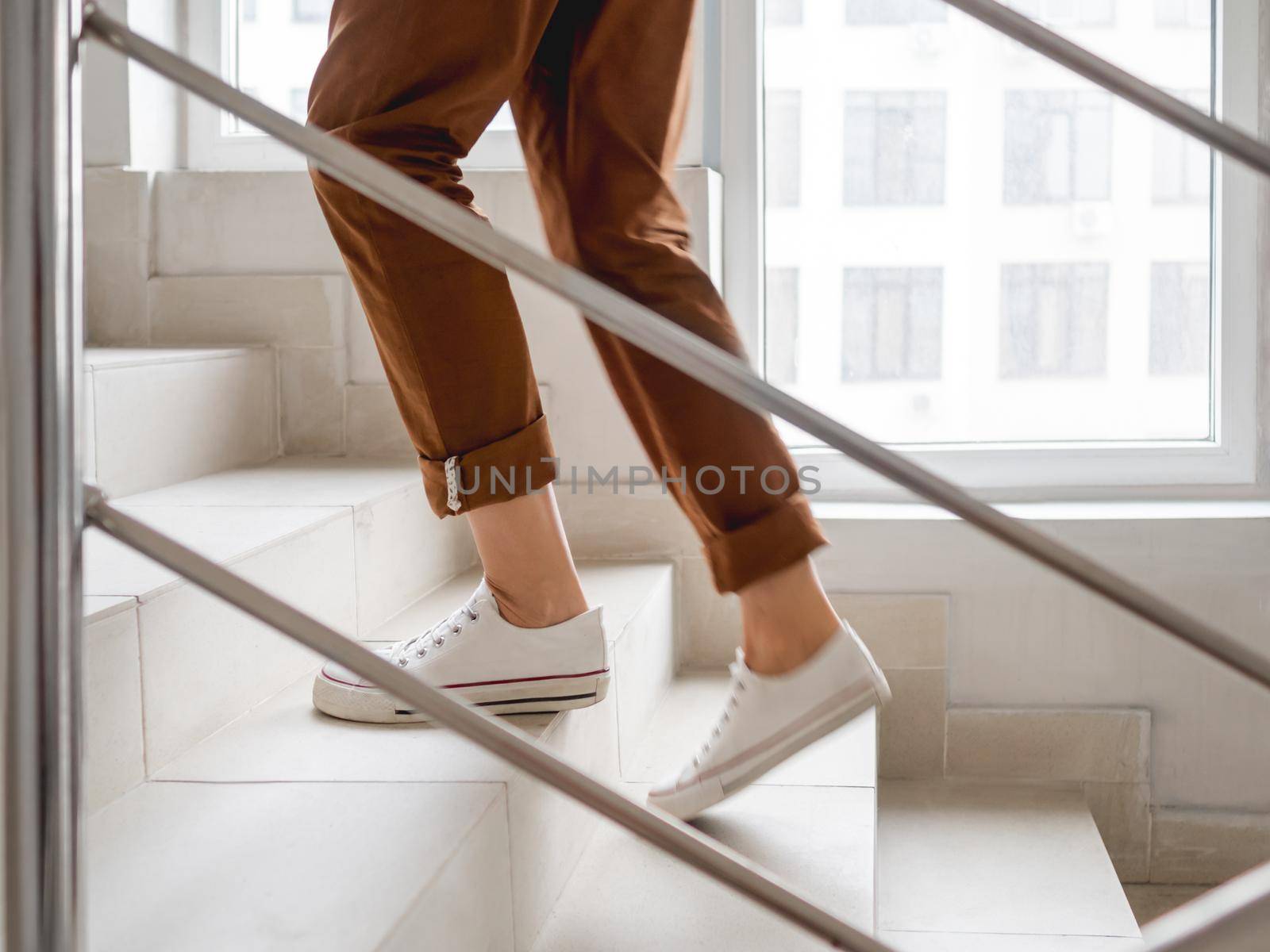 Woman in white sneakers and khaki trousers goes upstairs to her apartment. White staircase in apartment building. Casual outfit, urban fashion. Physical exercises. by aksenovko