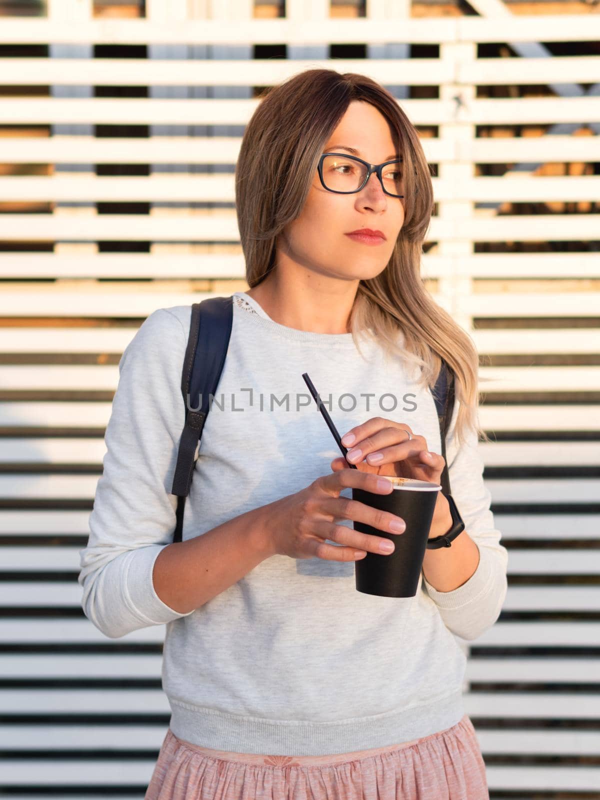 Woman with cup of coffee meets sunset on wooden pier. Female with curly hair and eyeglasses enjoys nature outdoors. Summer casual clothes. Summer vibes. by aksenovko