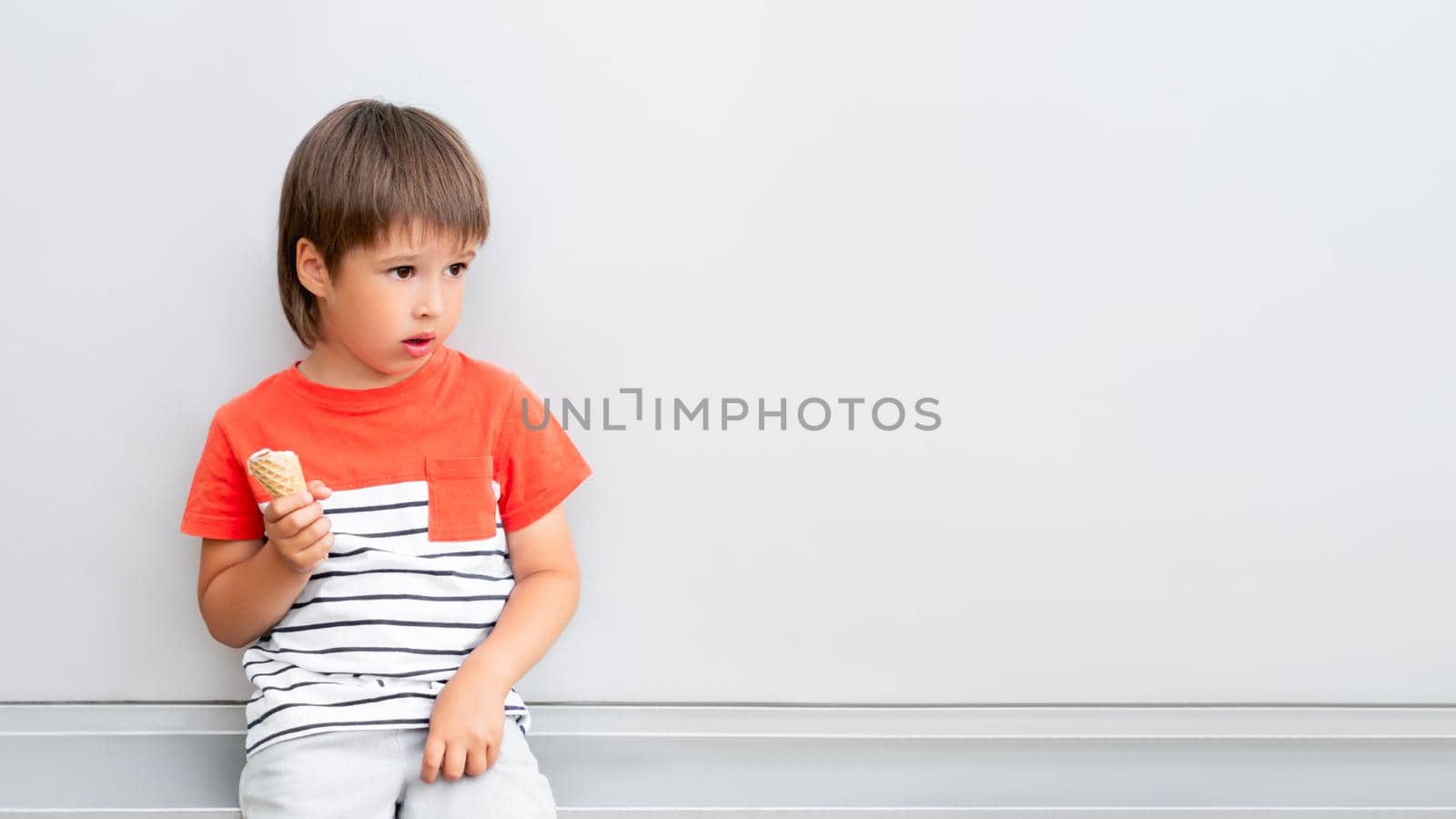 Little boy eats ice-cream in waffle cone. Child with cold dessert. Puzzled kid on grey background with copy space.