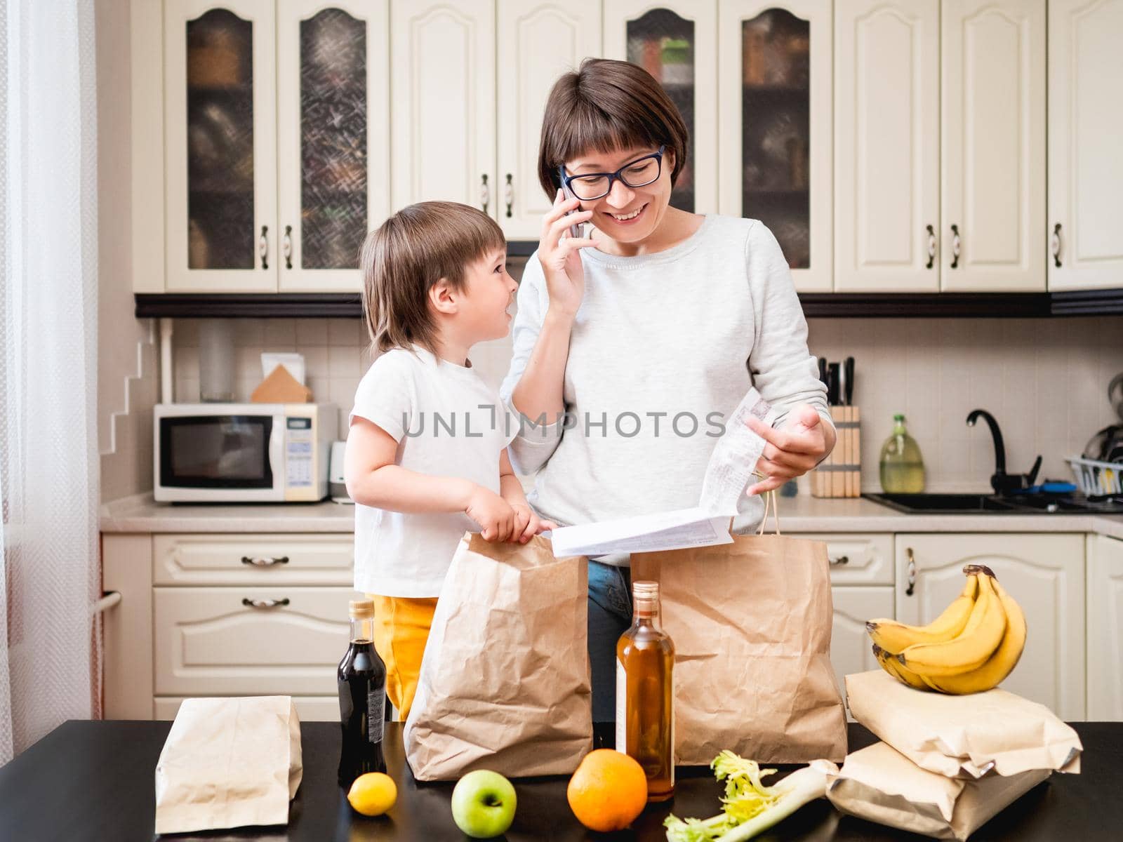 Woman and toddler boys sorts out purchases in the kitchen. Grocery delivery in paper bags. Online order from grocery store. Mother and son at kitchen. by aksenovko