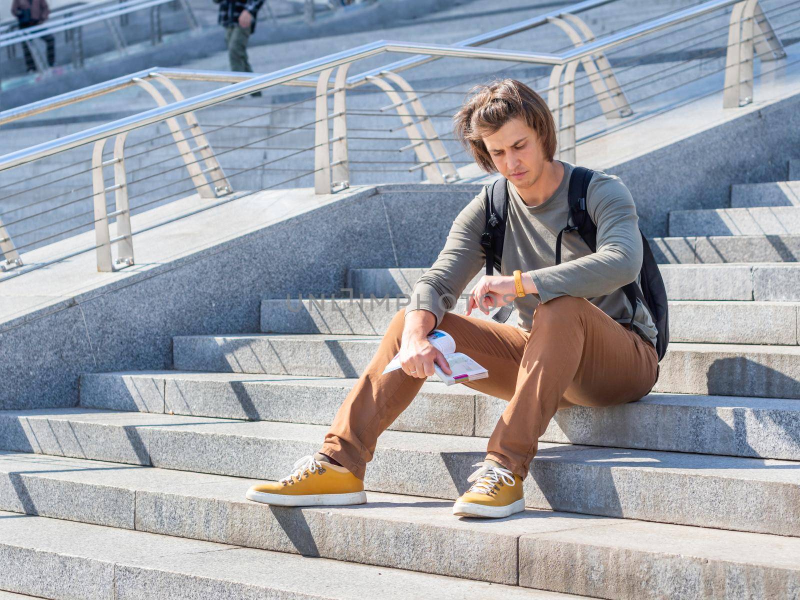 Man sits with book on stone staircase in park and waits for somebody. He looks on wrist-watch. Solo-travelling around city. Urban tourism.