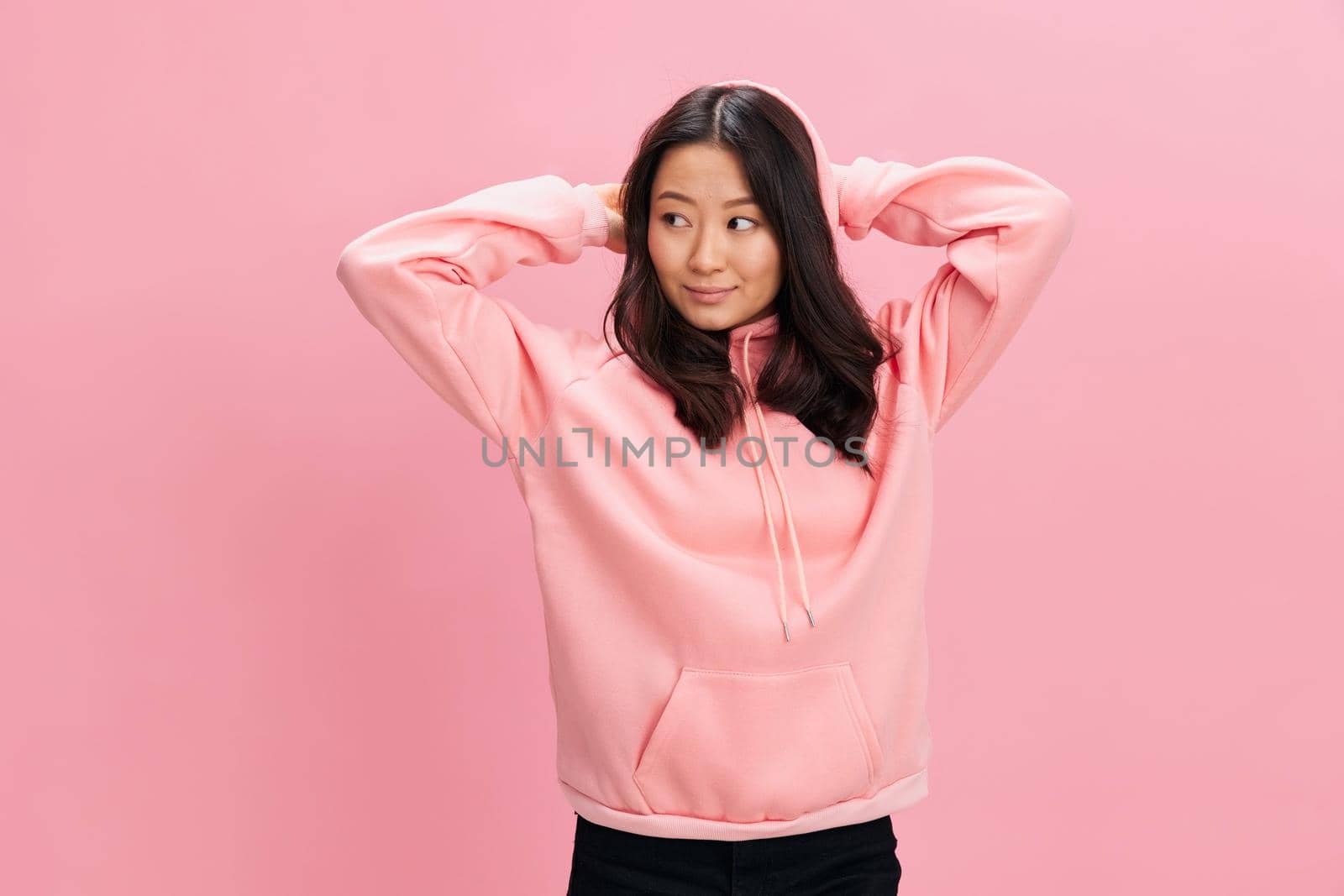 Smiling happy cute Asian student young lady in pink hoodie sweatshirt hold hands behind head posing isolated on over pink studio background. Good offer. Fashion New Collection concept