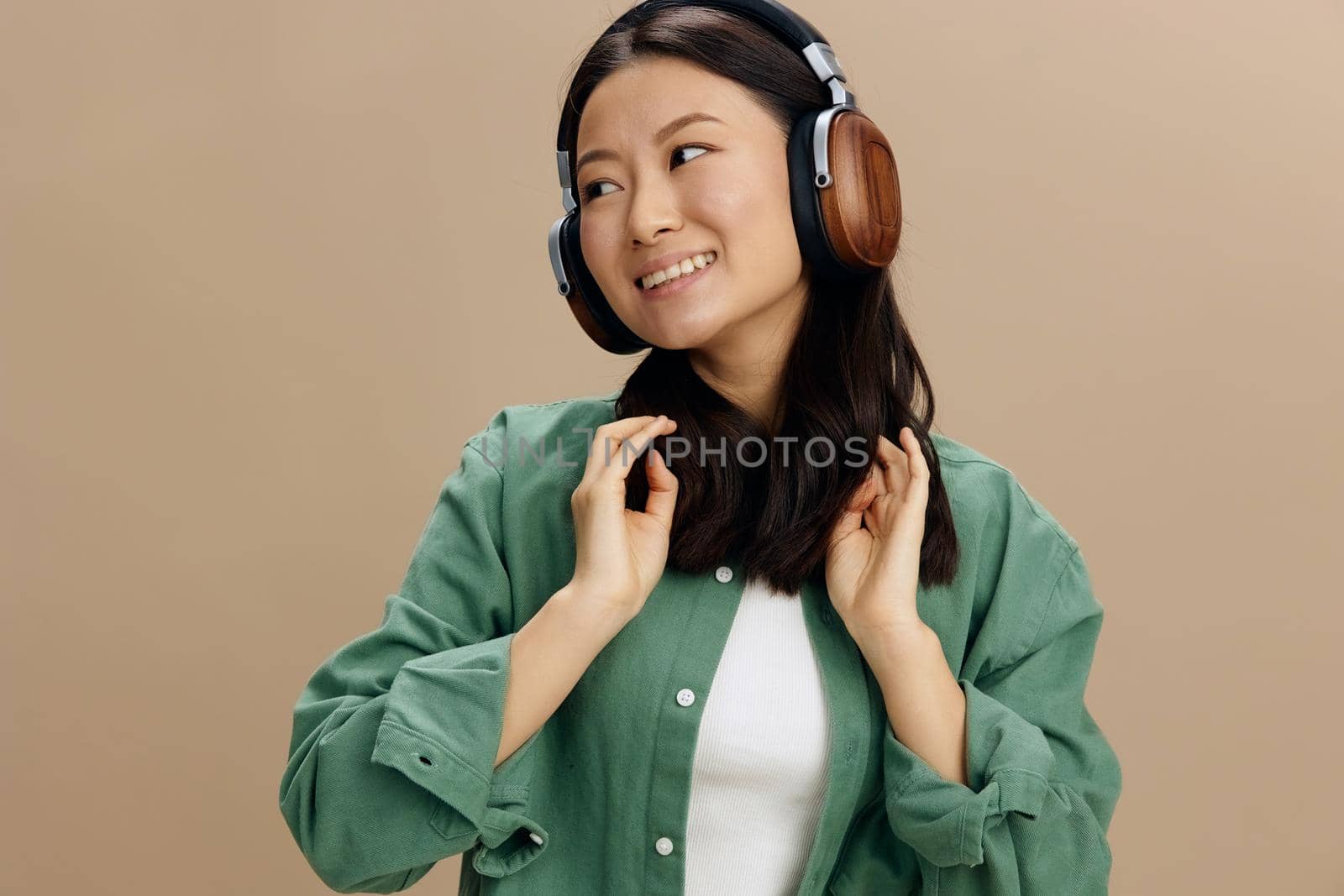 Enjoy it. Happy cute Asian student young woman in khaki green shirt touch headphones look aside posing isolated on beige pastel studio background. Cool fashion offer. Music App Platform Ad concept