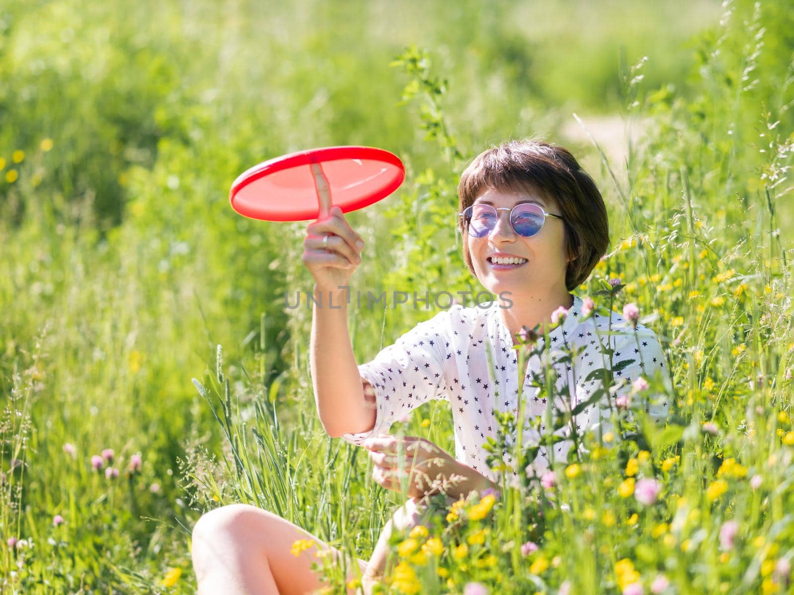 Woman in colorful sunglasses plays with red frisbee, enjoys sunlight and flower fragrance on grass field. Summer vibes. Relax outdoors. Self-soothing. by aksenovko