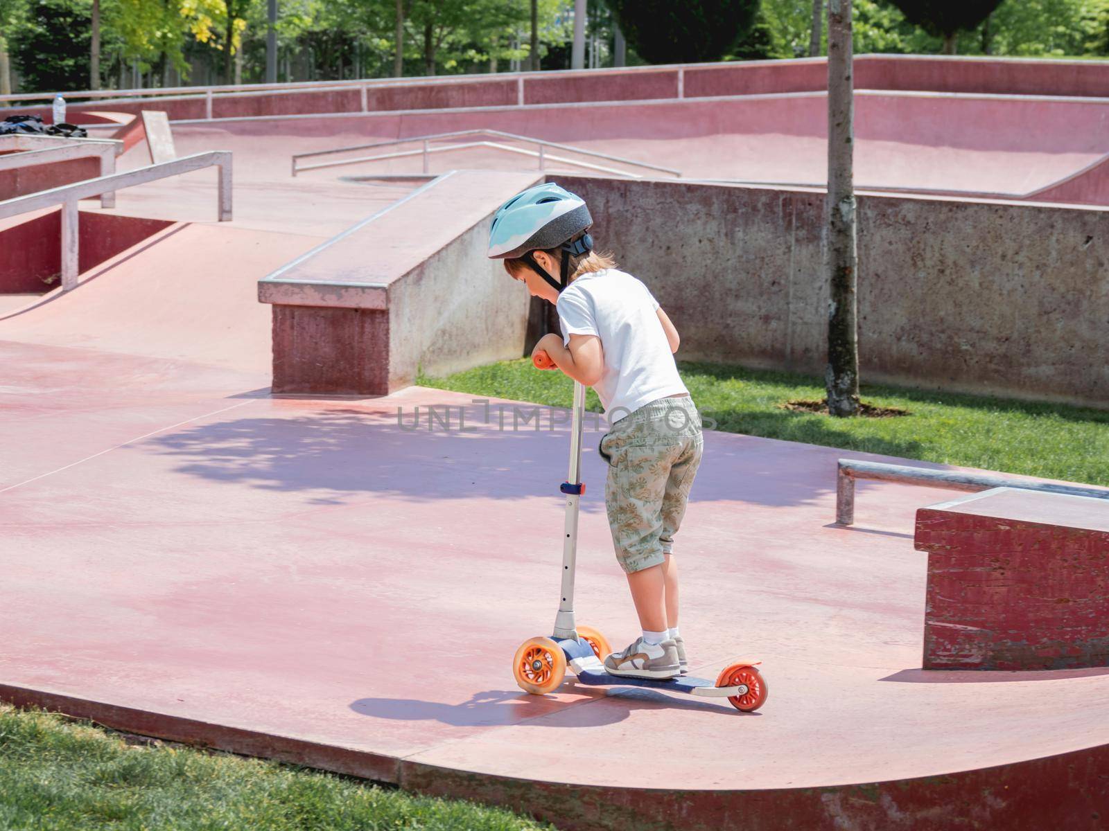 Little boy rides kick scooter in skate park. Special concrete bowl structures in urban park. Training to skate at summer. by aksenovko