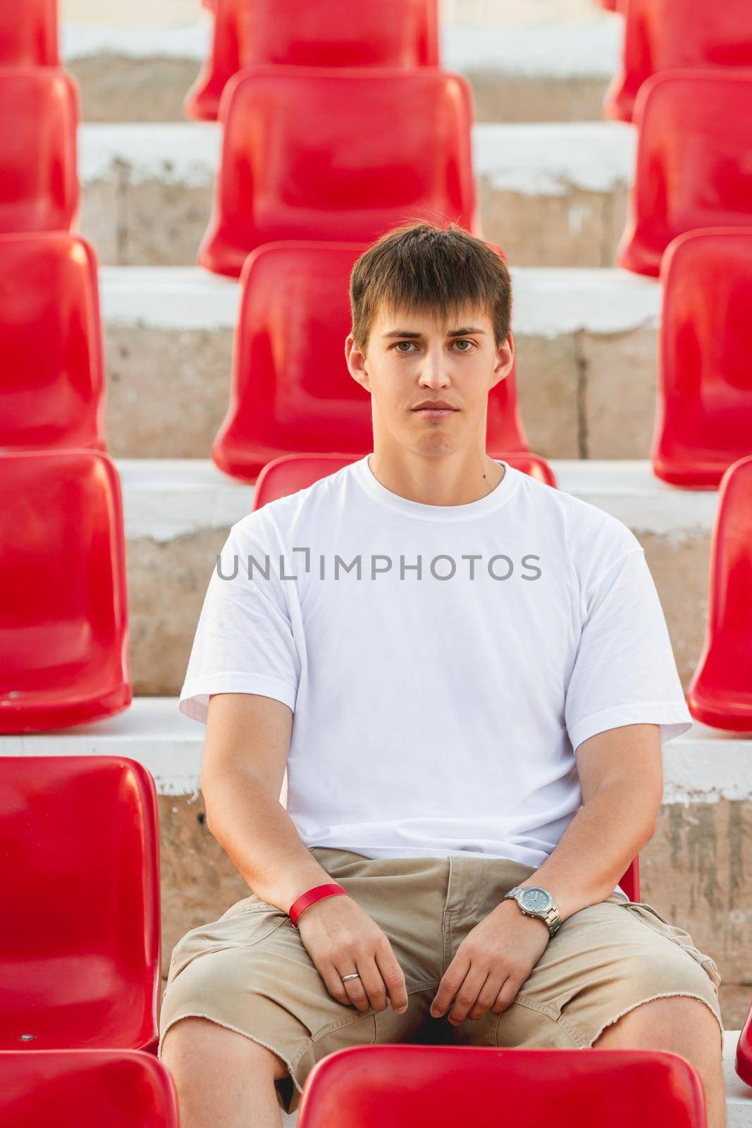 Bored young man sitting in deserted audience with bright red seats. Absence of people in open-air amphitheater due to coronavirus COVID-19 pandemic lockdown and quarantine.